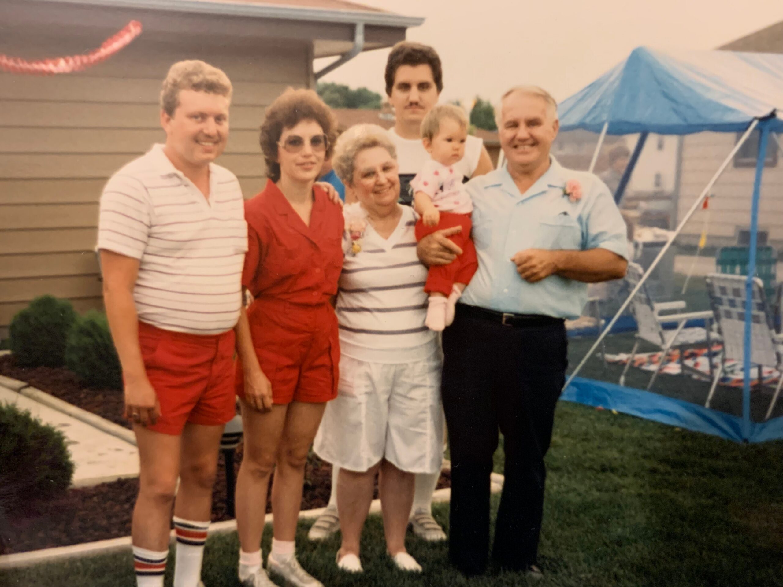 Family posing together in a backyard, with a toddler held by an older woman and a blue canopy trampoline in the background.