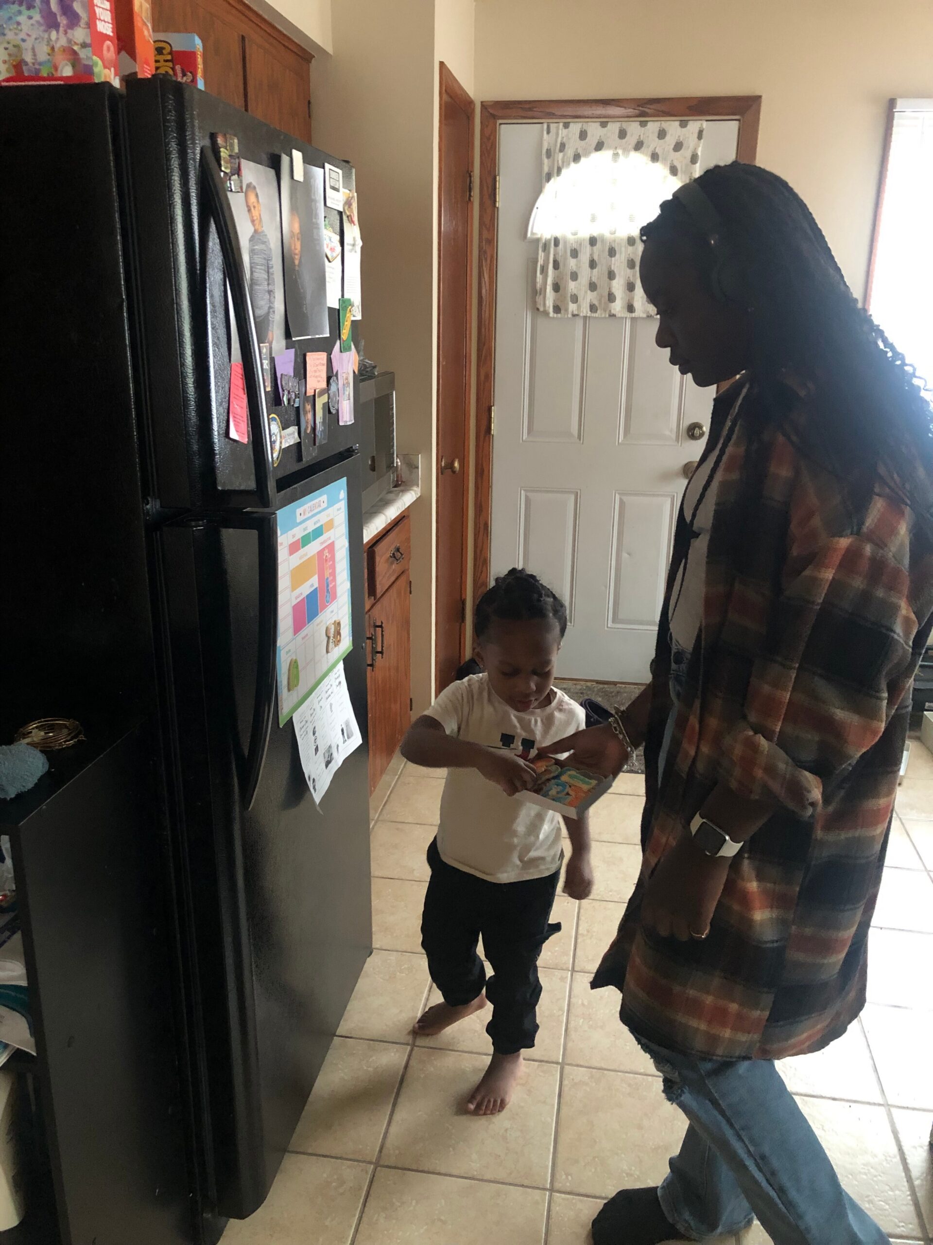 Woman with braided hair and a plaid shirt stands beside a child, showing and reading a colorful magazine in a kitchen by a magnet-filled refrigerator.
