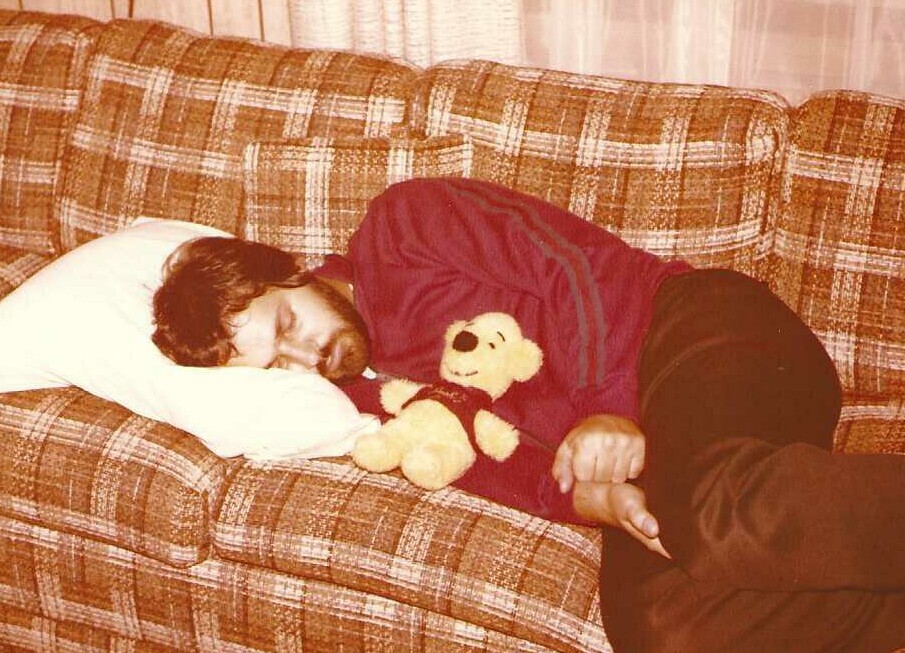 Man sleeping on a plaid sofa holding a teddy bear under his arm.