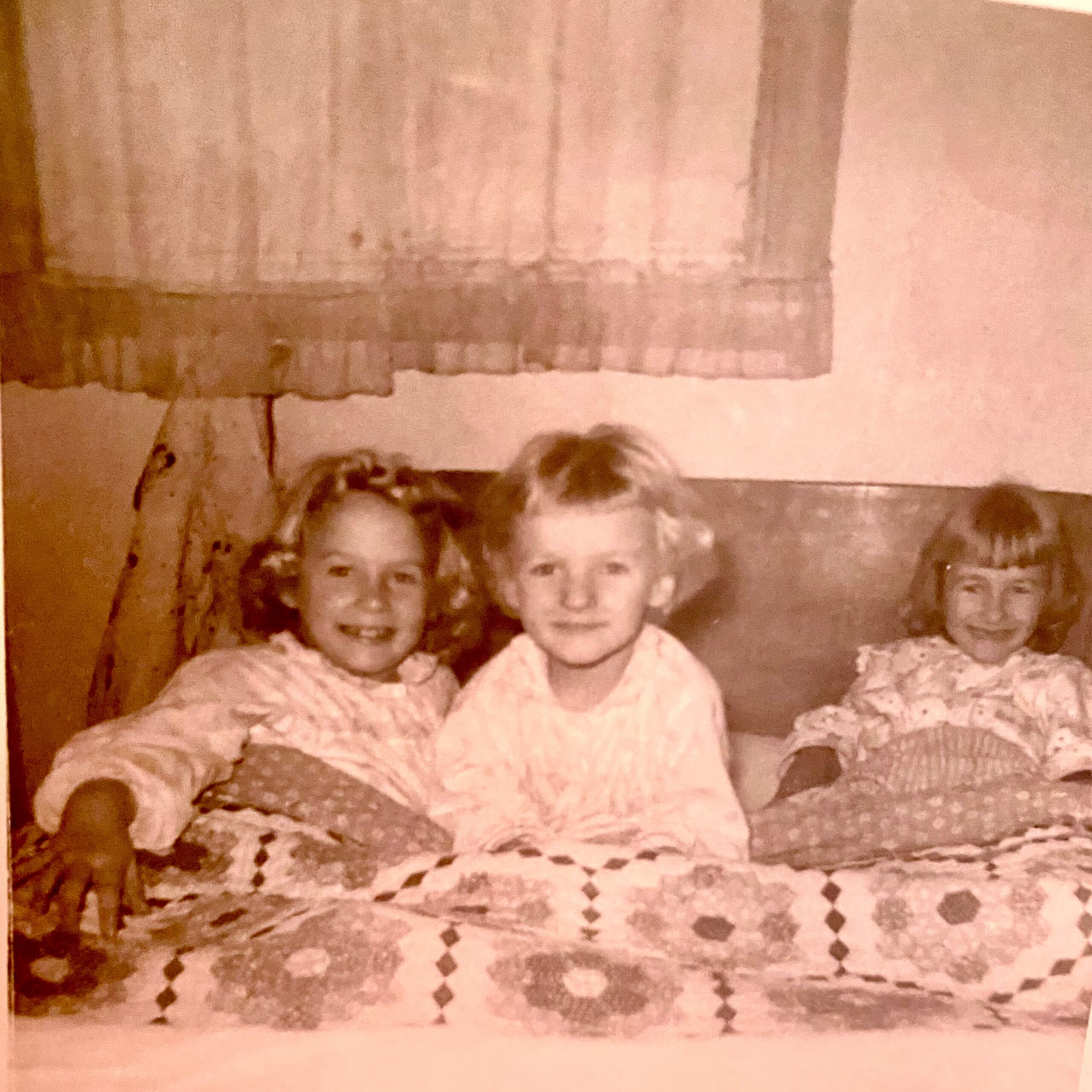 Old sepia-toned family photo of six young children sitting together on a crocheted blanket, smiling at the camera.
