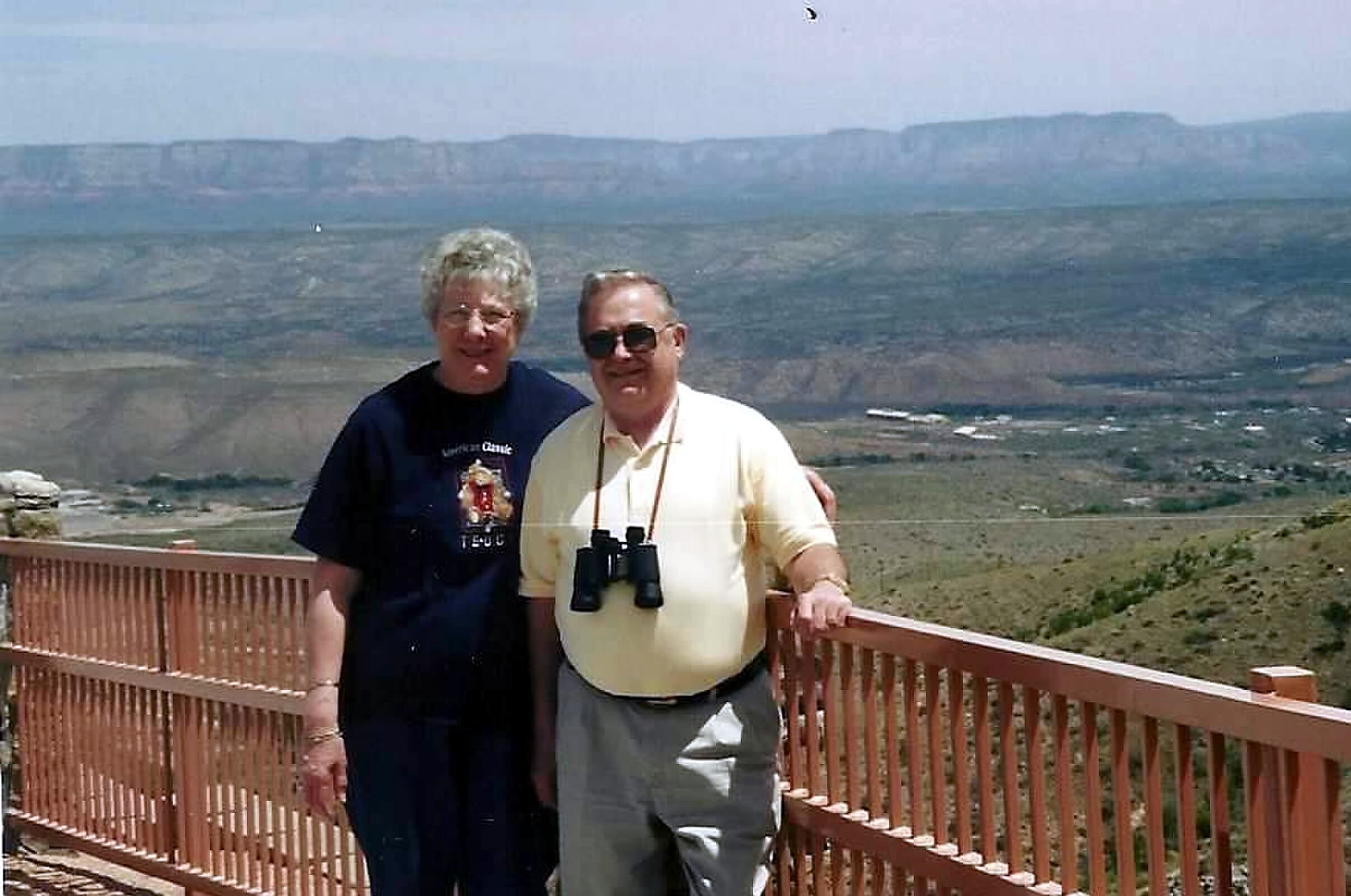 Two men standing at a railing overlook with a desert valley and distant mesas behind them; one in a dark shirt, the other in a light shirt with binoculars. (Informative)