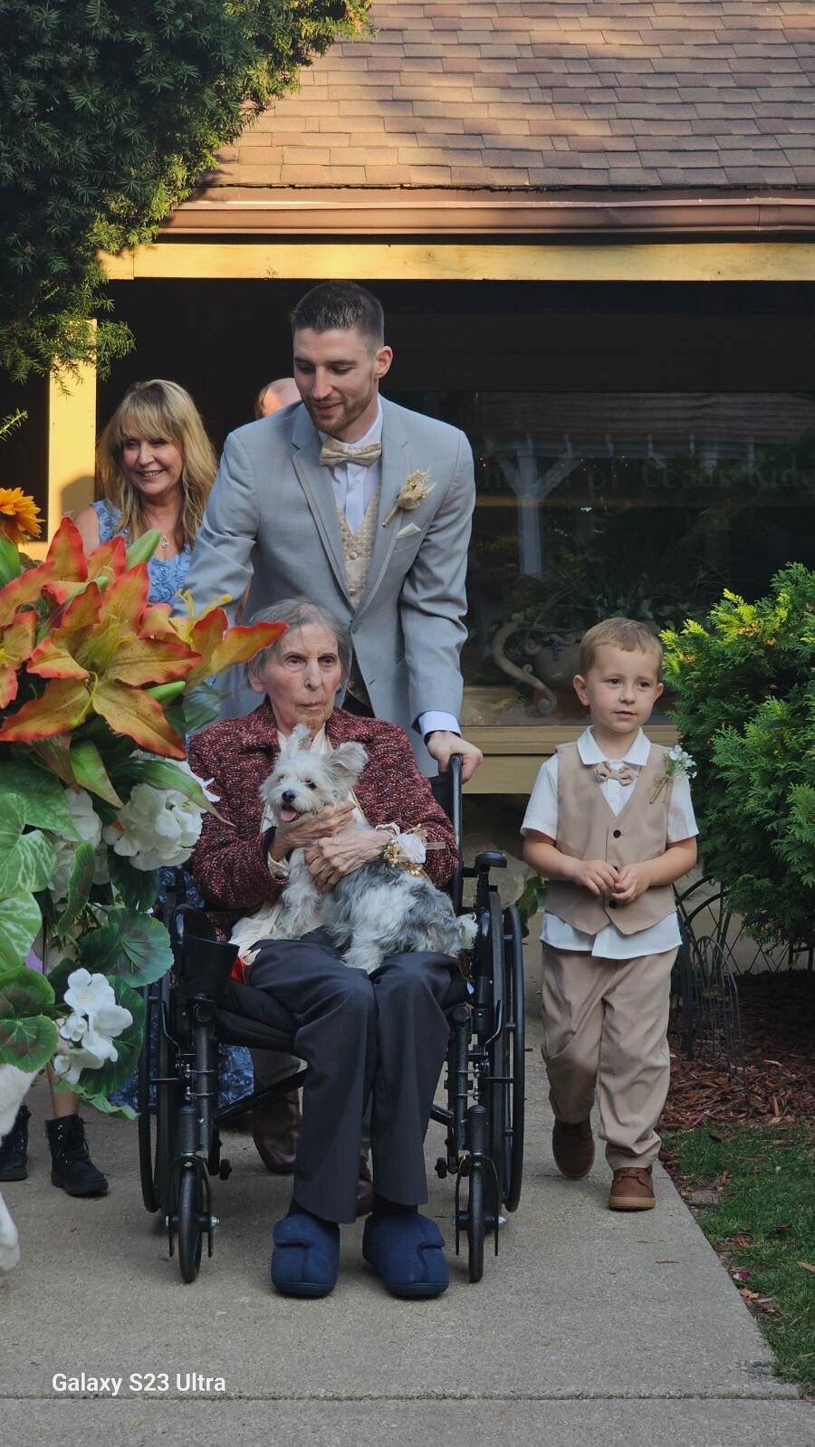 Older woman in a wheelchair holding a small white dog, with a man in a light gray suit pushing her and a young boy in beige formal wear nearby, outside a home with colorful flowers behind them.