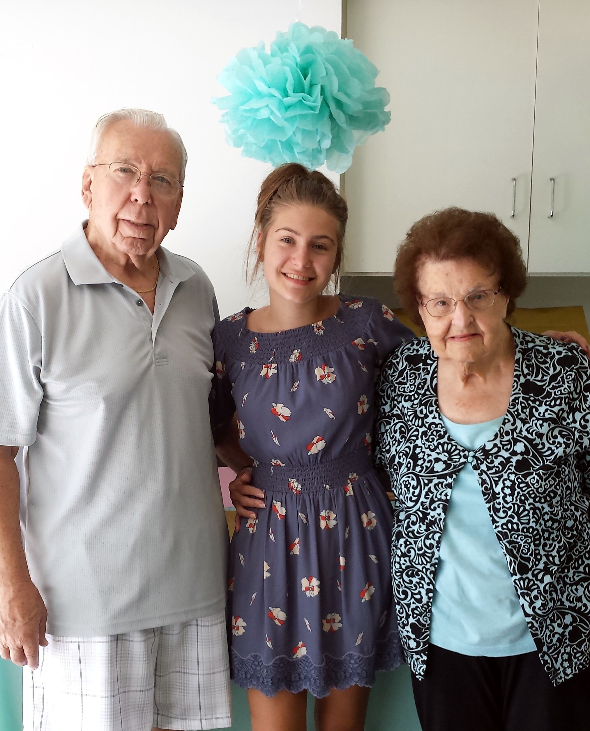 Three generations posing indoors: an elderly man, a young woman, and an elderly woman with a large turquoise tissue paper pom-pom overhead.