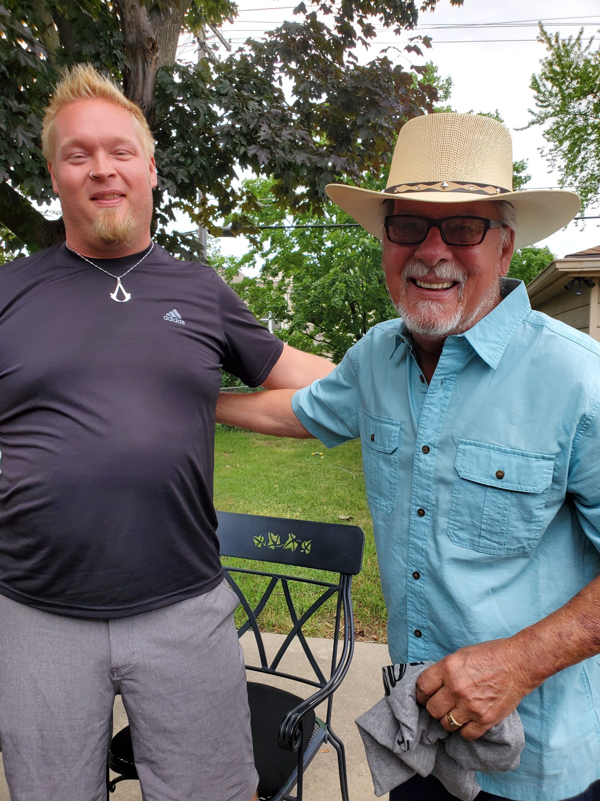 Two men posing outdoors, smiling for the photo; one in a black shirt with a necklace, the other wearing a straw hat, sunglasses, and a light blue button-up shirt.