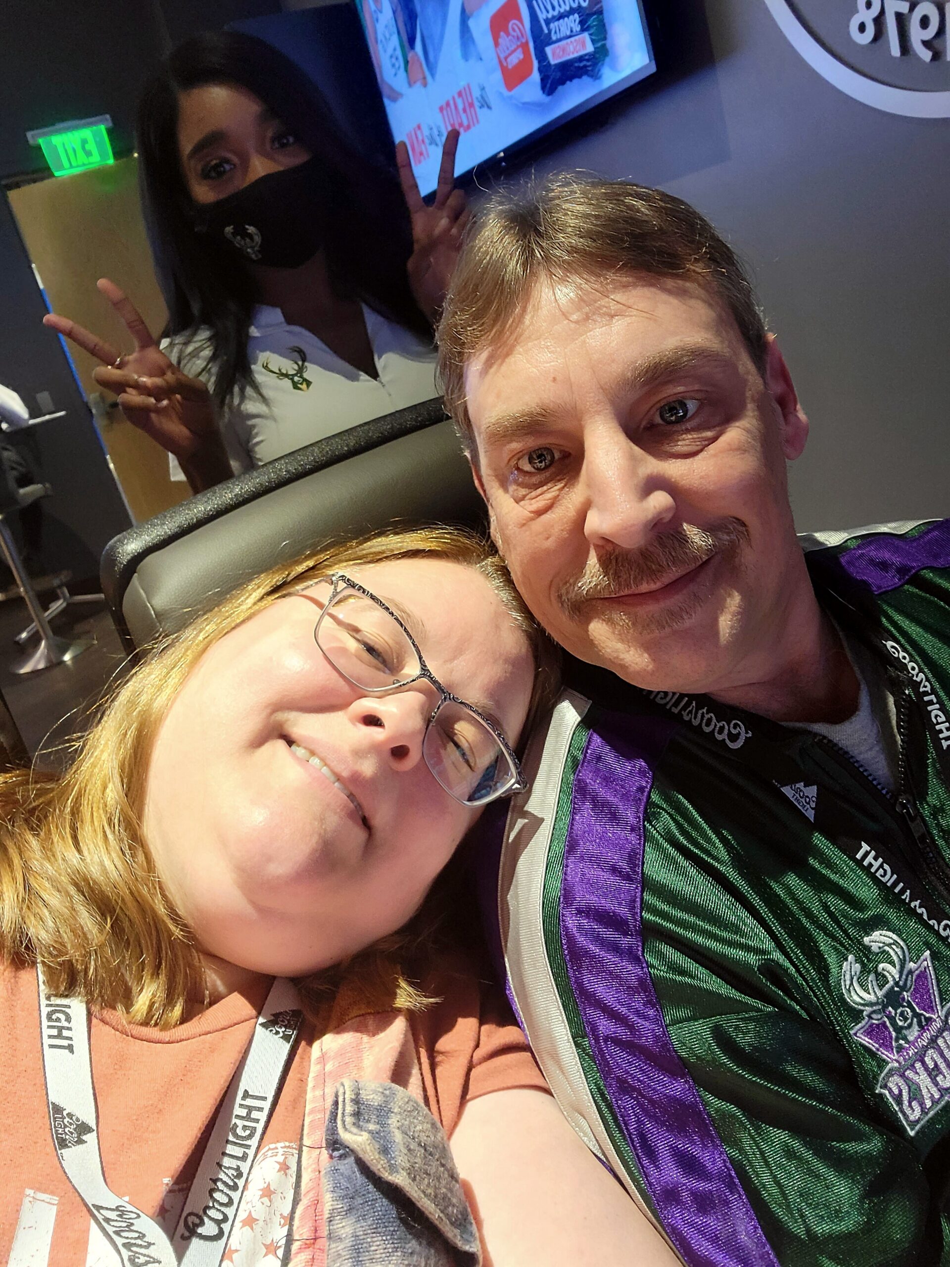 Close-up selfie of a smiling man and woman resting their heads together, with the woman wearing a black face mask and giving a peace sign in the background behind them.
