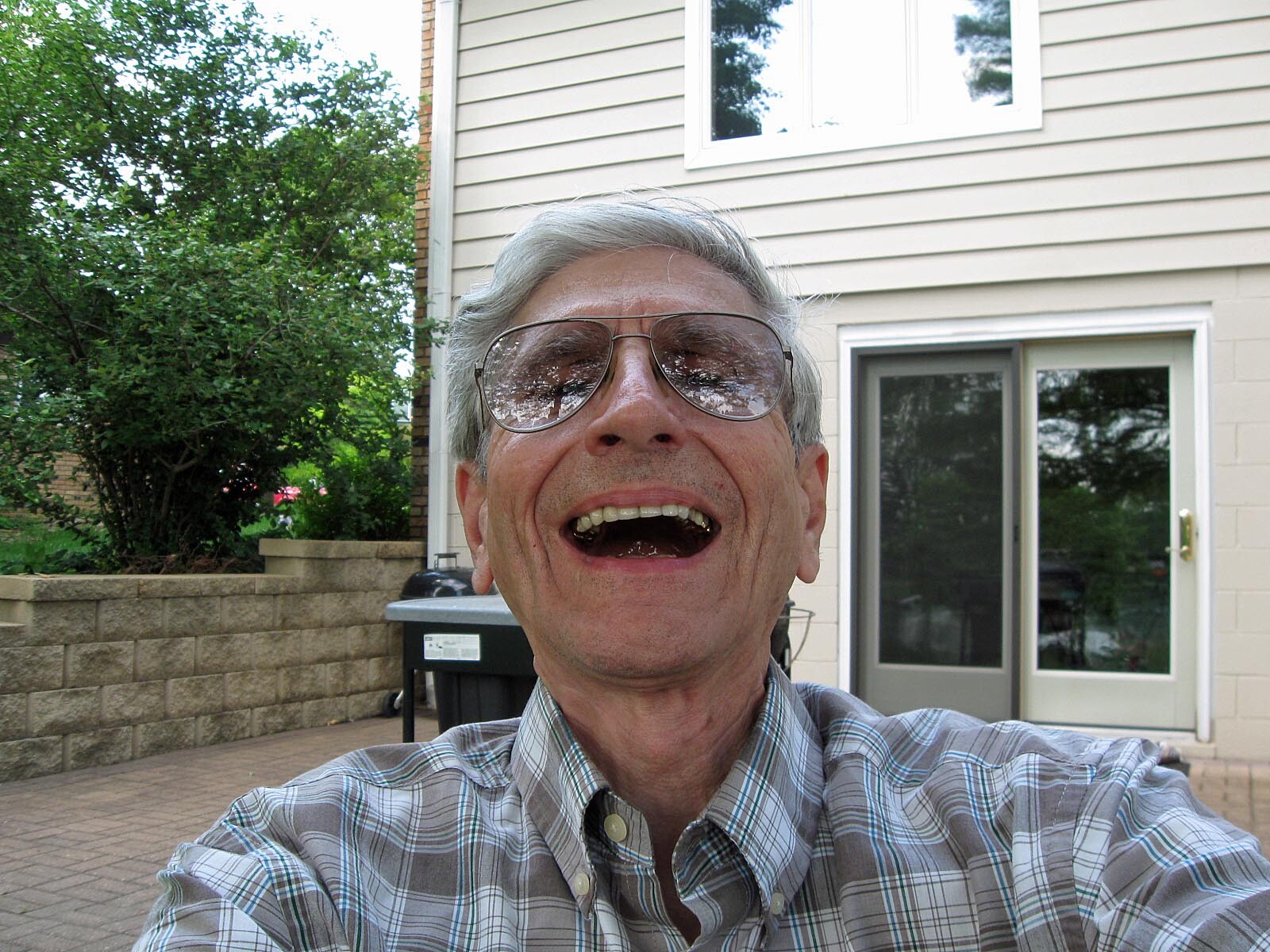 Smiling older man wearing glasses and a plaid shirt takes a selfie in a backyard with a beige house, trees, and a sliding glass door behind him.