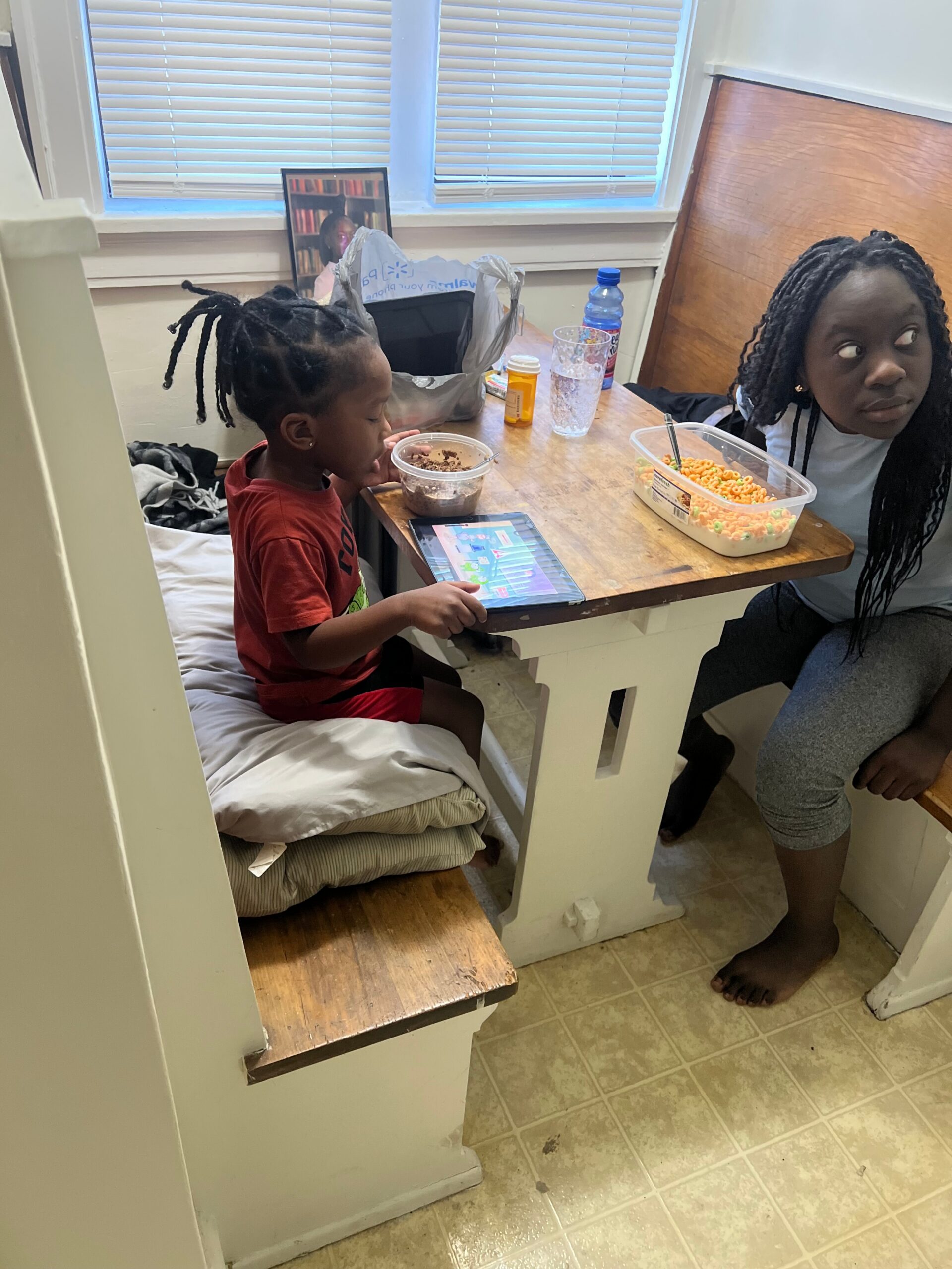 Two children eat breakfast at a small wooden table; one has a cereal bowl and tablet nearby, with a framed photo in the background.