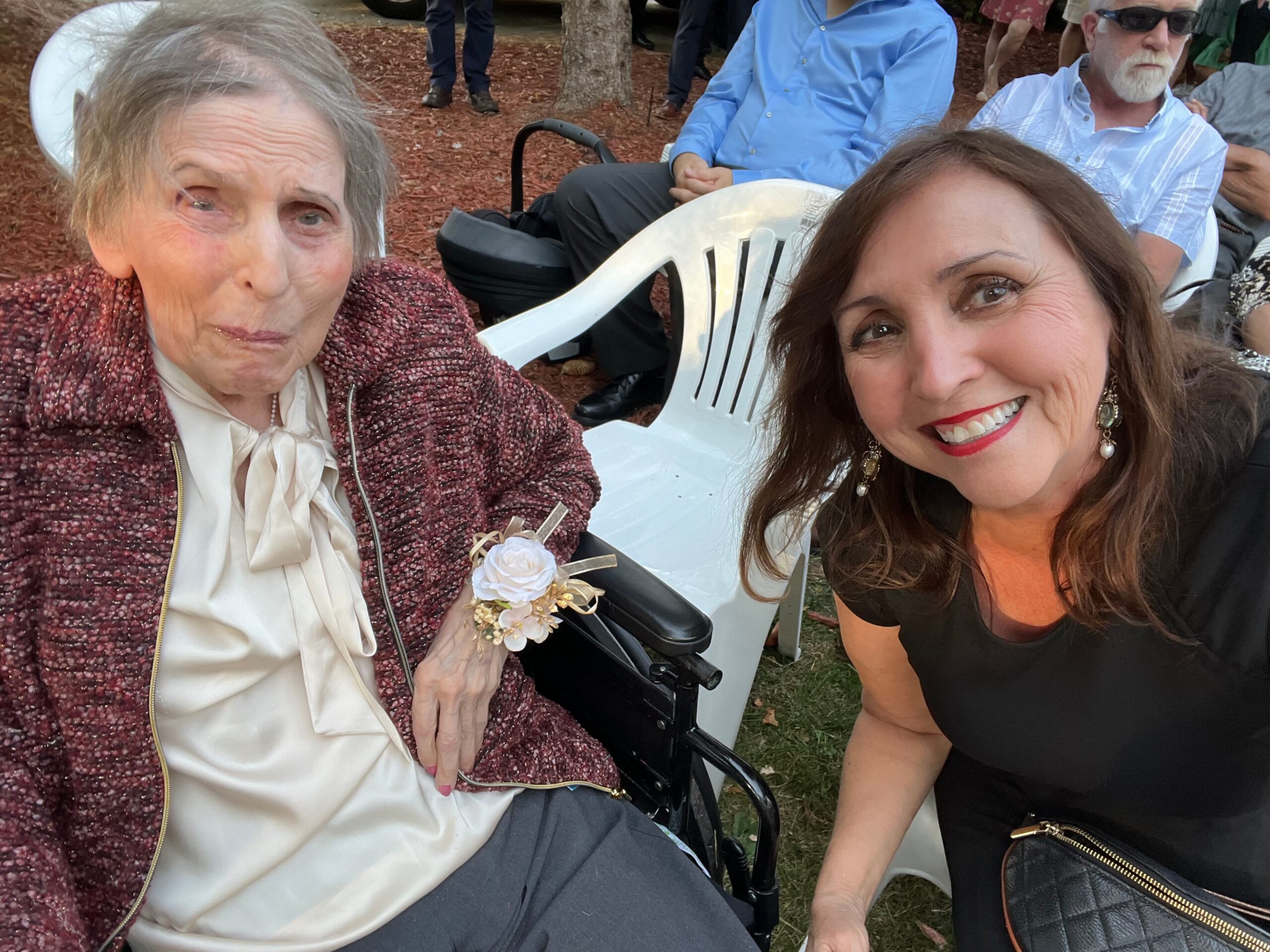 Smiling woman in black dress takes a selfie with an elderly woman in a wheelchair outdoors at a gathering.