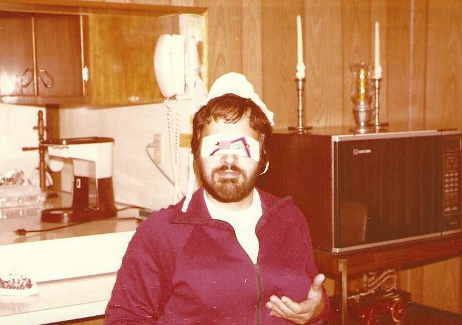 Man with a beard sits in a retro kitchen, wearing a white head covering and a makeshift blindfold over his eyes, with a microwave and coffee maker in the background.