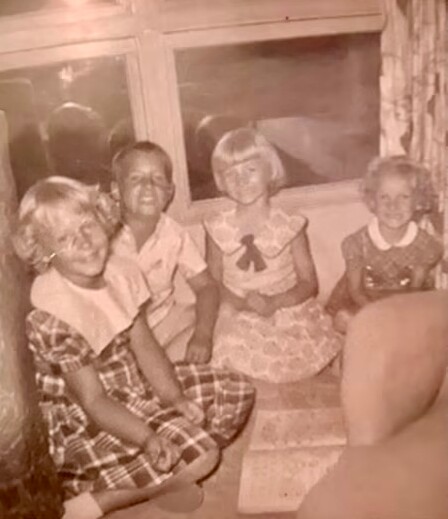 Four children sit on the floor by a window in a sepia-toned vintage photo, wearing dresses and shirts.