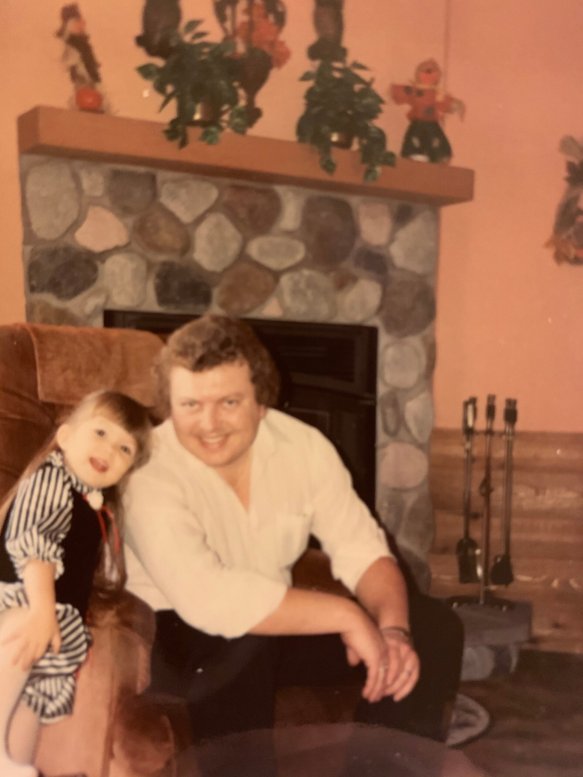 Man in a white shirt sits next to a young girl in a striped dress on a sofa in front of a stone fireplace with a wooden mantel and holiday decorations above it.