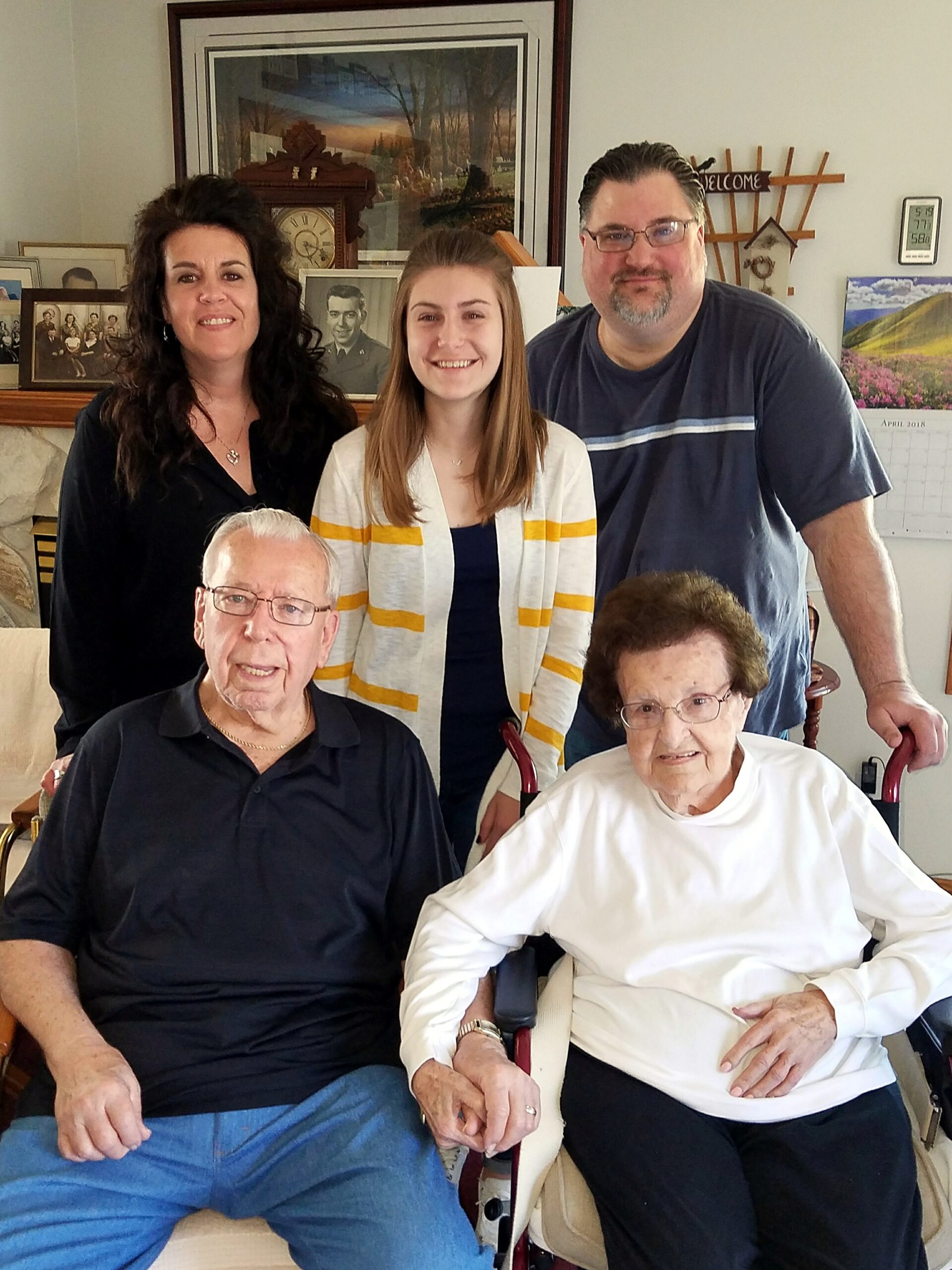 Five-family group posing in a living room: two seniors seated in front and three adults standing behind them, with photos on the mantel behind.