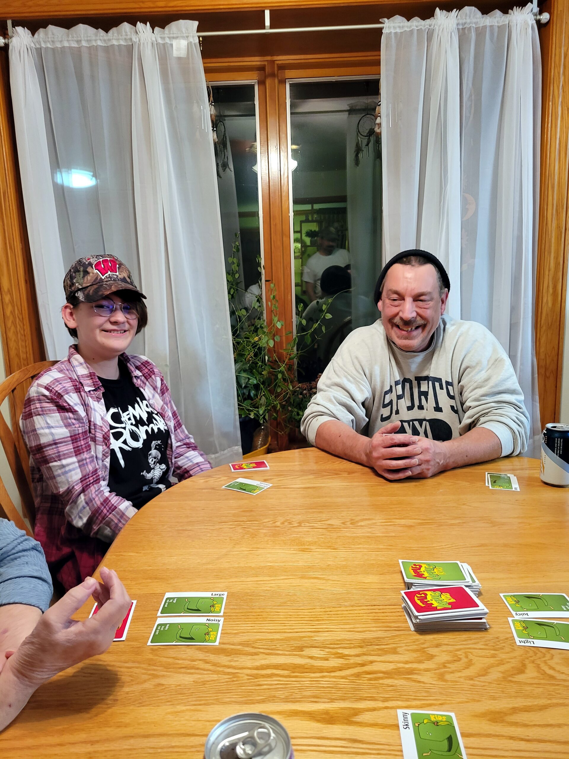 Two people sit at a wooden table, playing a card game; green-backed cards are spread out, a can on the table behind them.