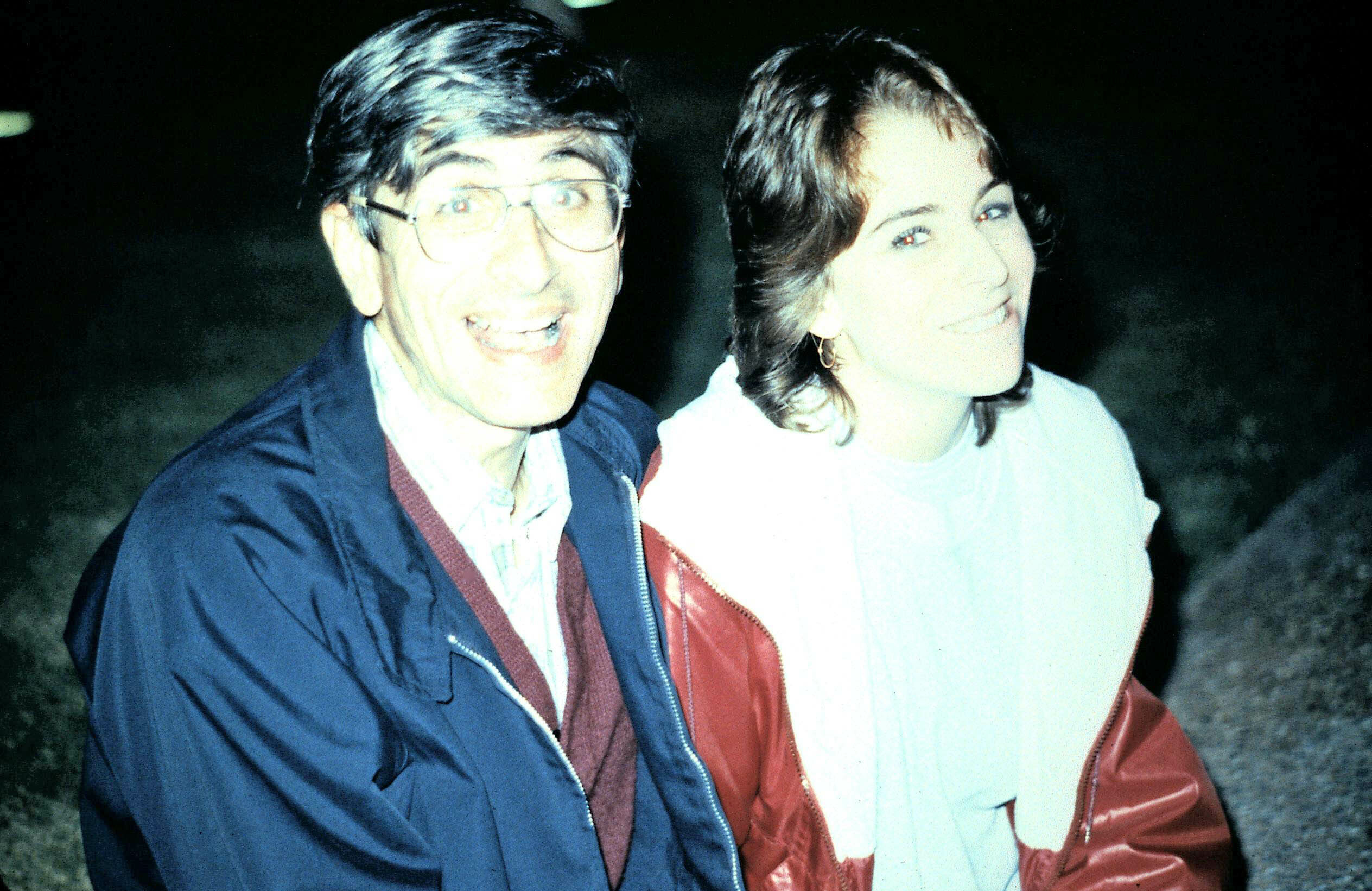 Two friends pose for a nighttime outdoor photo, smiling while wearing jackets (man on left, woman on right).