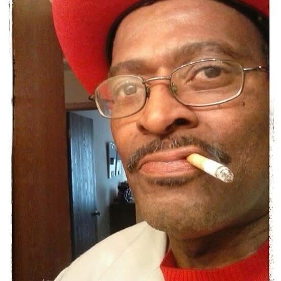 Close-up of a man wearing glasses and a red hat, smoking a cigarette indoors.