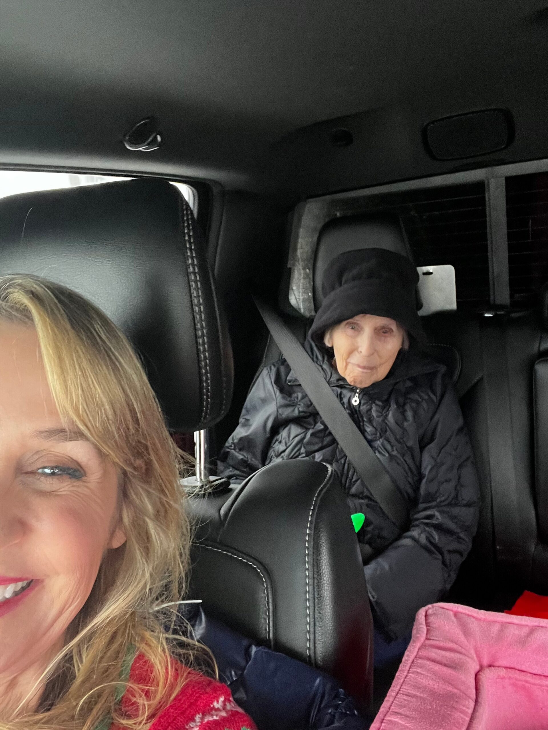 Smiling woman with blond hair in the front seat of a car, with an elderly woman wearing a black hat in the back seat, both wearing jackets and seat belts.