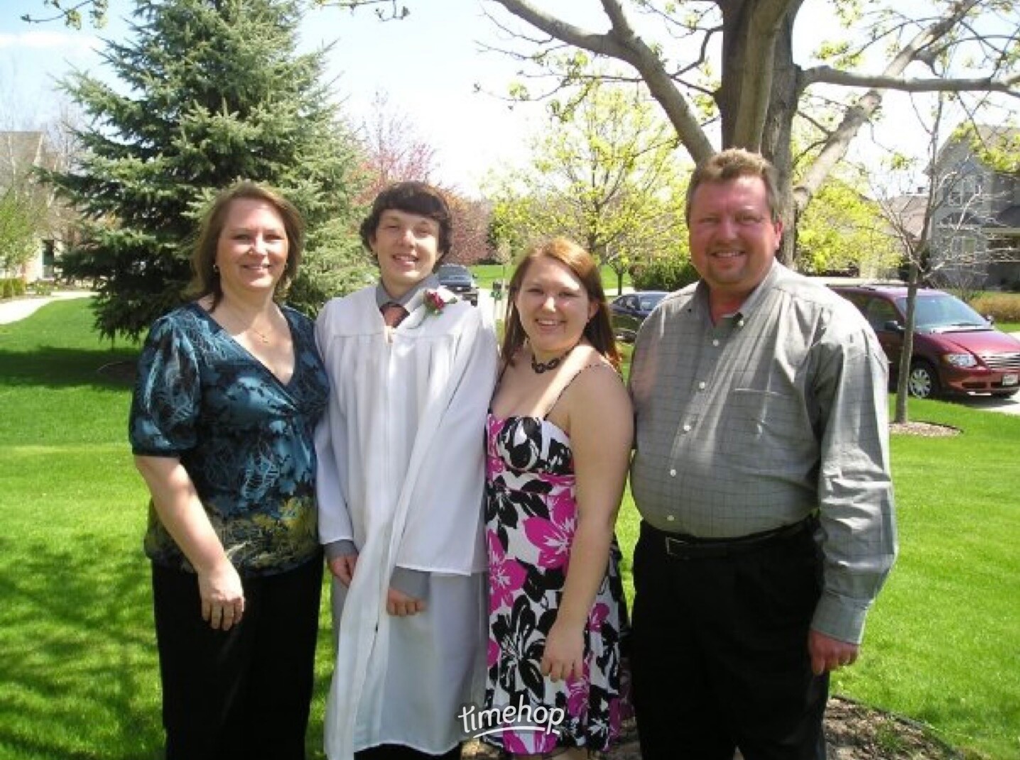 Four people smiling outdoors on a sunny day; a graduate in a white gown stands second from left, flanked by two adults and a friend or relative in casual attire on a green lawn.