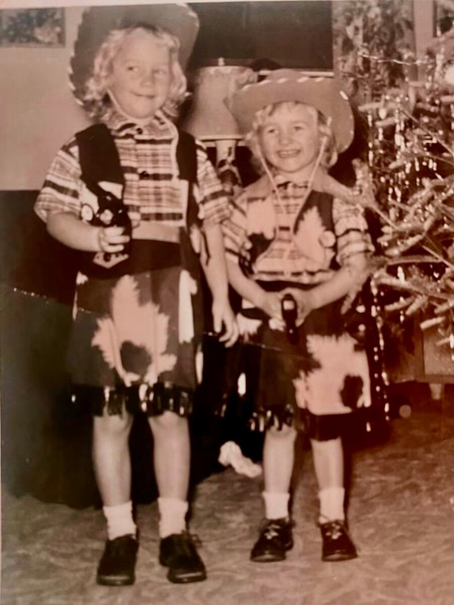 Two children in cowboy costumes pose indoors beside a Christmas tree, one holding a toy horse.