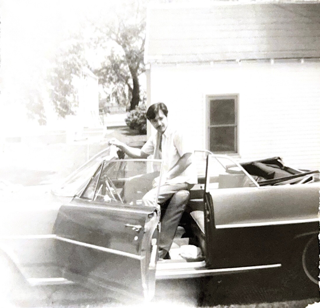 Man in a light shirt and tie entering the driver's seat of a dark convertible with the door open.