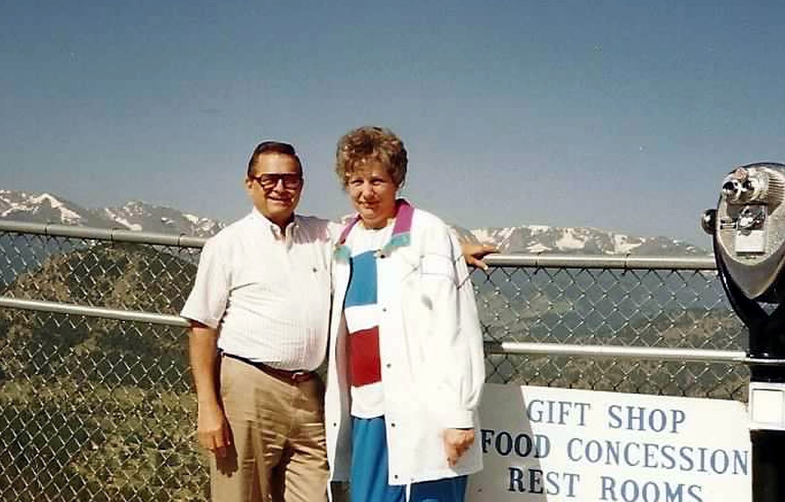 Two people stand together in front of a chain-link fence with snow-capped mountains in the background.