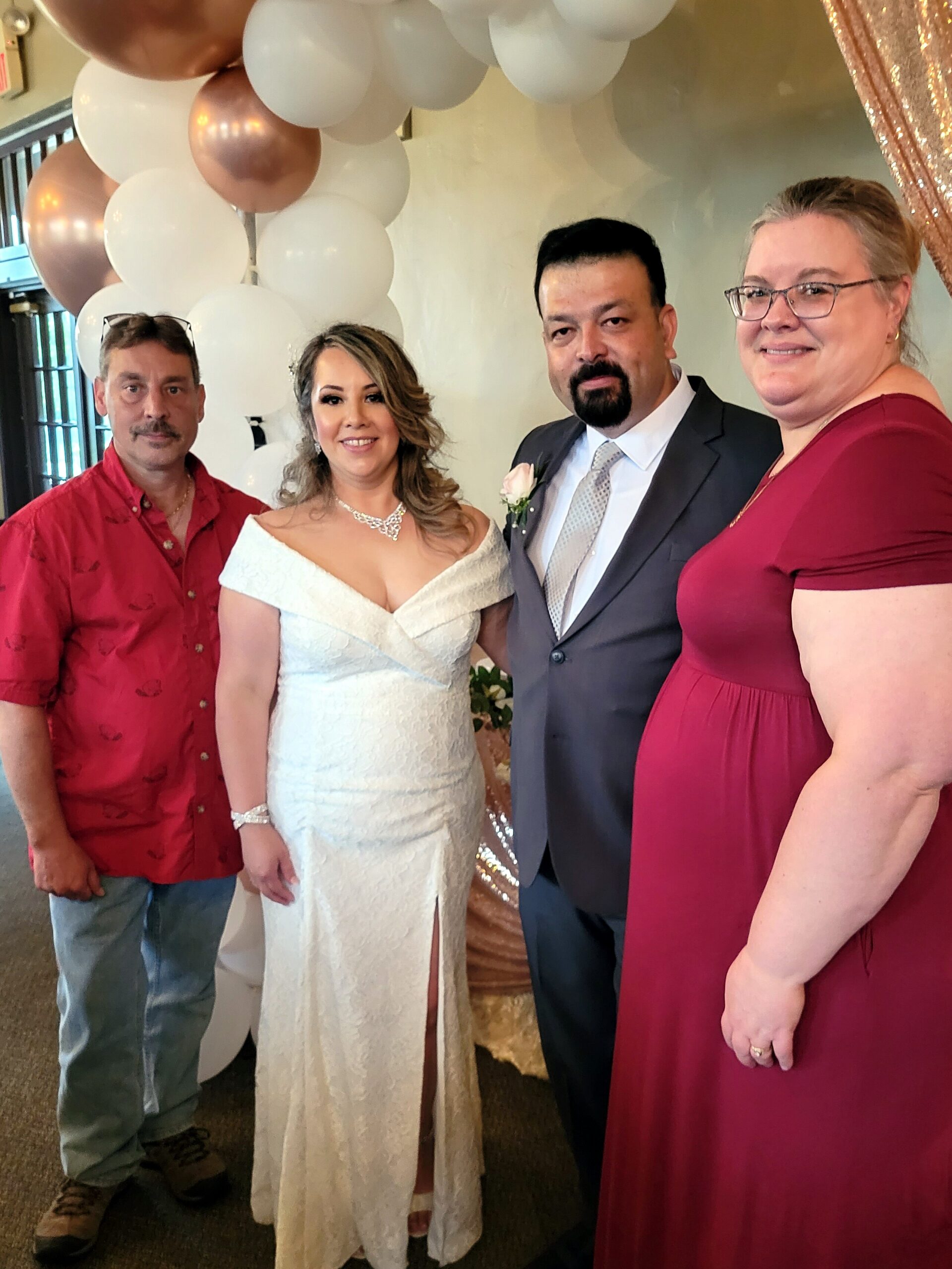 Bride in a white lace gown and her groom in a gray suit pose with two guests under a balloon arch, smiling at a wedding event.