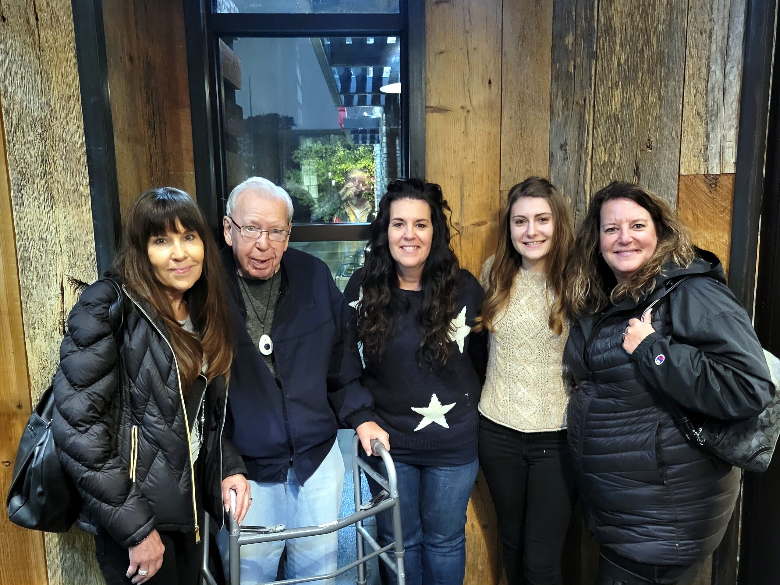 Five adults standing close together, smiling for a group photo in a wood-paneled indoor space with a metal walker visible in front of them.
