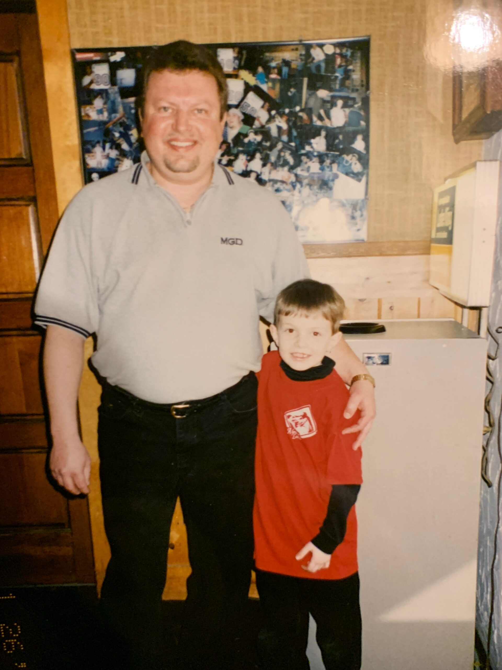 Smiling man with short hair and a goatee standing beside a young boy in a red shirt indoors, near a collage on the wall.