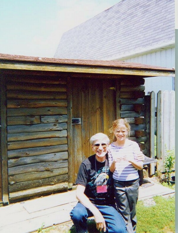 Two people pose together outdoors in front of a rustic log shed, smiling at the camera, on a sunny day.
