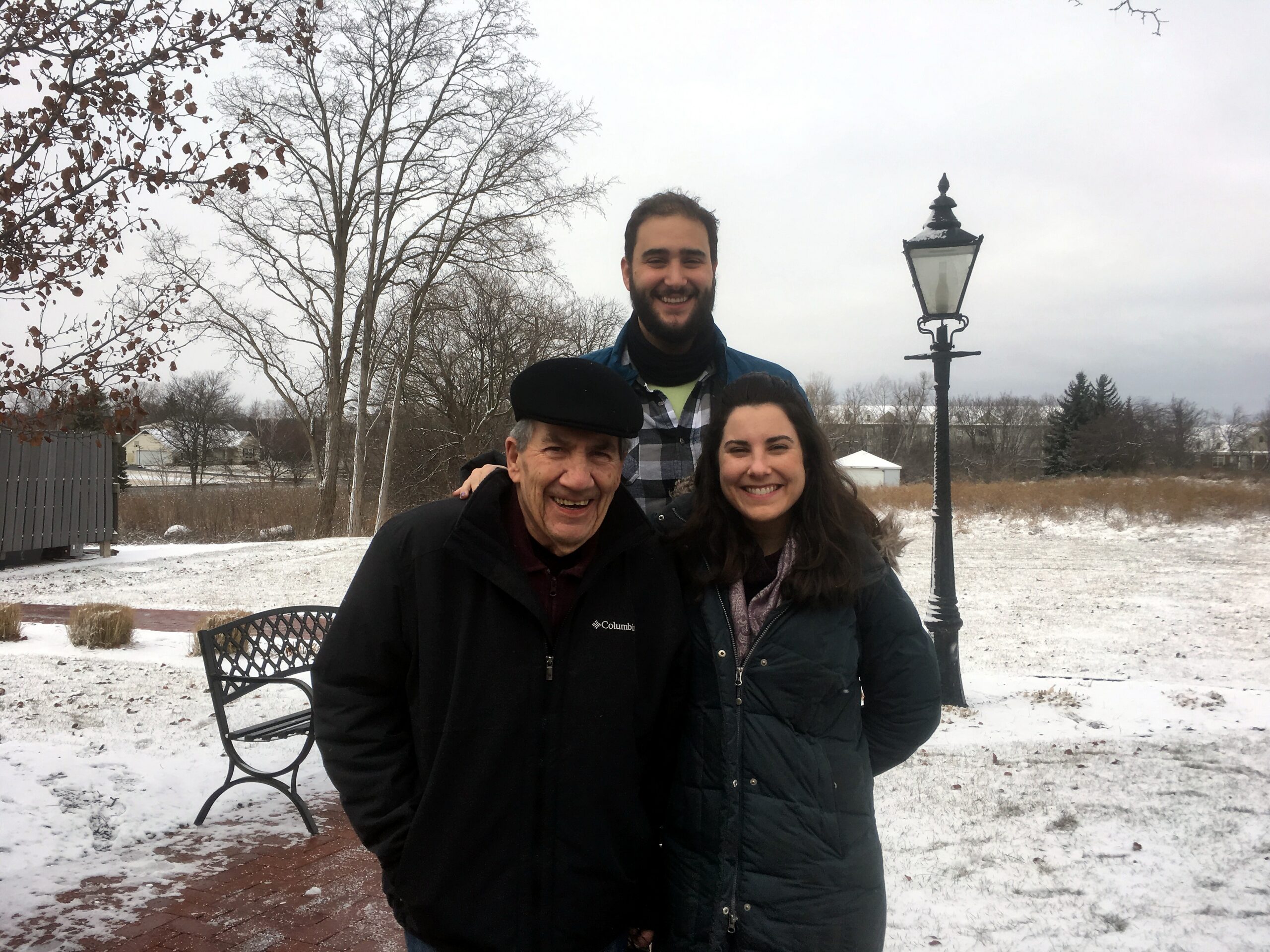 Three people in winter coats smile together outside on a snowy day, with bare trees and a decorative street lamp in the background after a snowfall.
