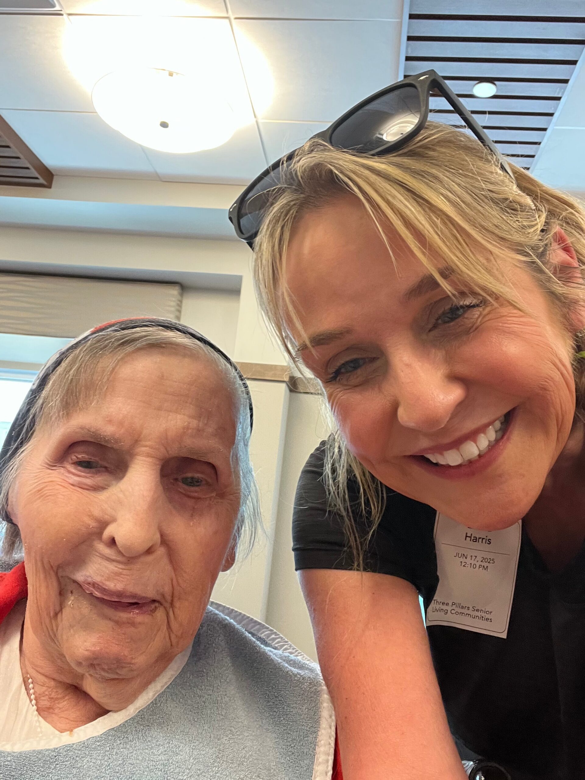 Close-up selfie of an elderly woman and a smiling younger woman in a bright indoor room, the younger with sunglasses on her head and a name badge.