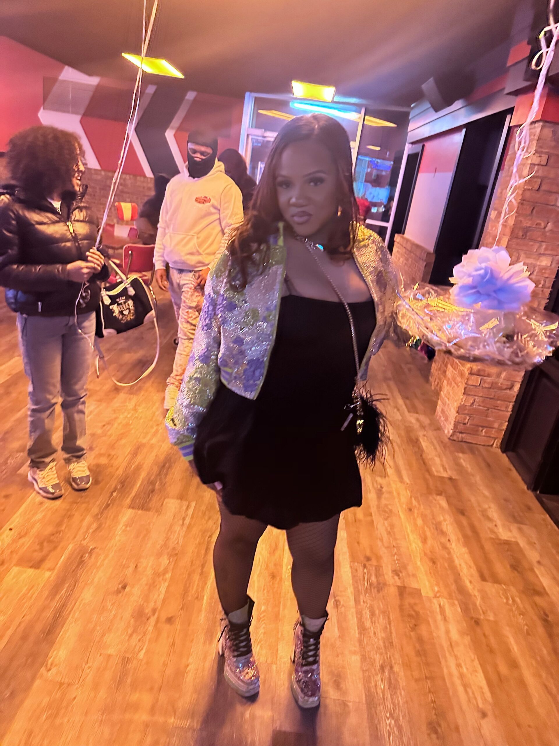 Young woman in a glittery jacket posing with a wrapped bouquet in a neon-lit room; people in the background.