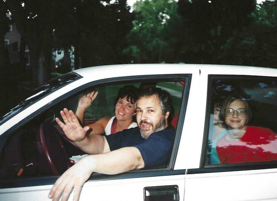 Three people ride in a white car; the man in the front seat waves at the camera while two women sit in the back, smiling.