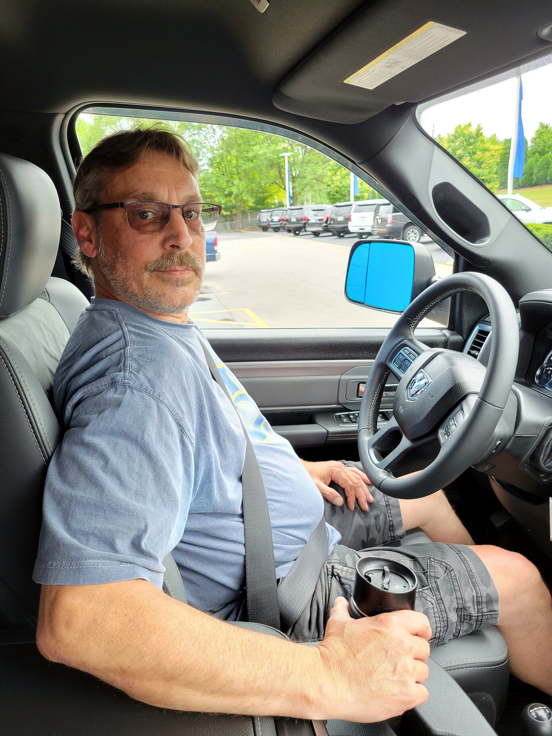 Man wearing glasses sits in the driver's seat of a pickup, buckled in with a seat belt, looking toward the camera inside a car interior.