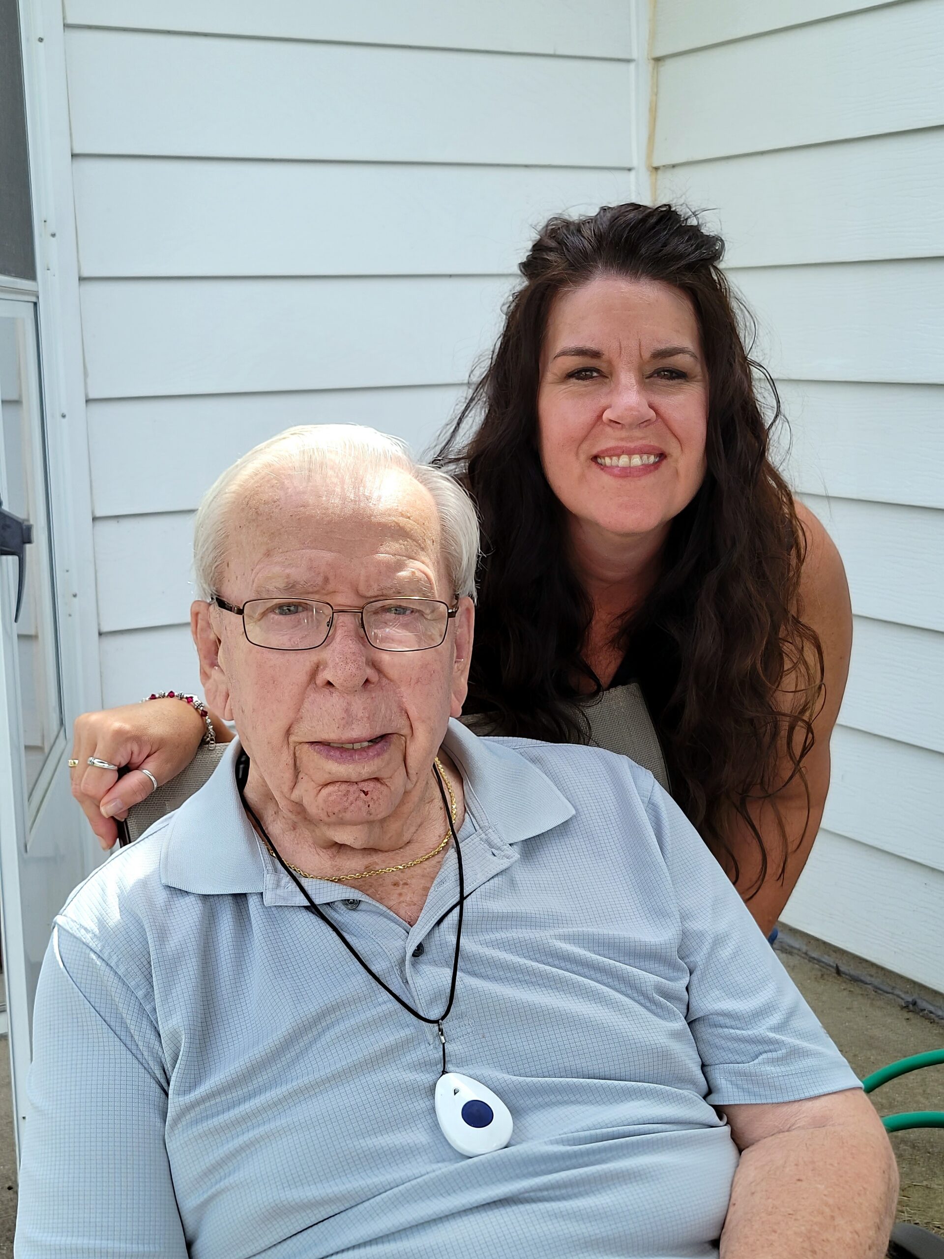 Smiling elderly man in a light blue shirt wearing glasses seated on a porch chair, with a younger woman with long dark hair smiling behind him.