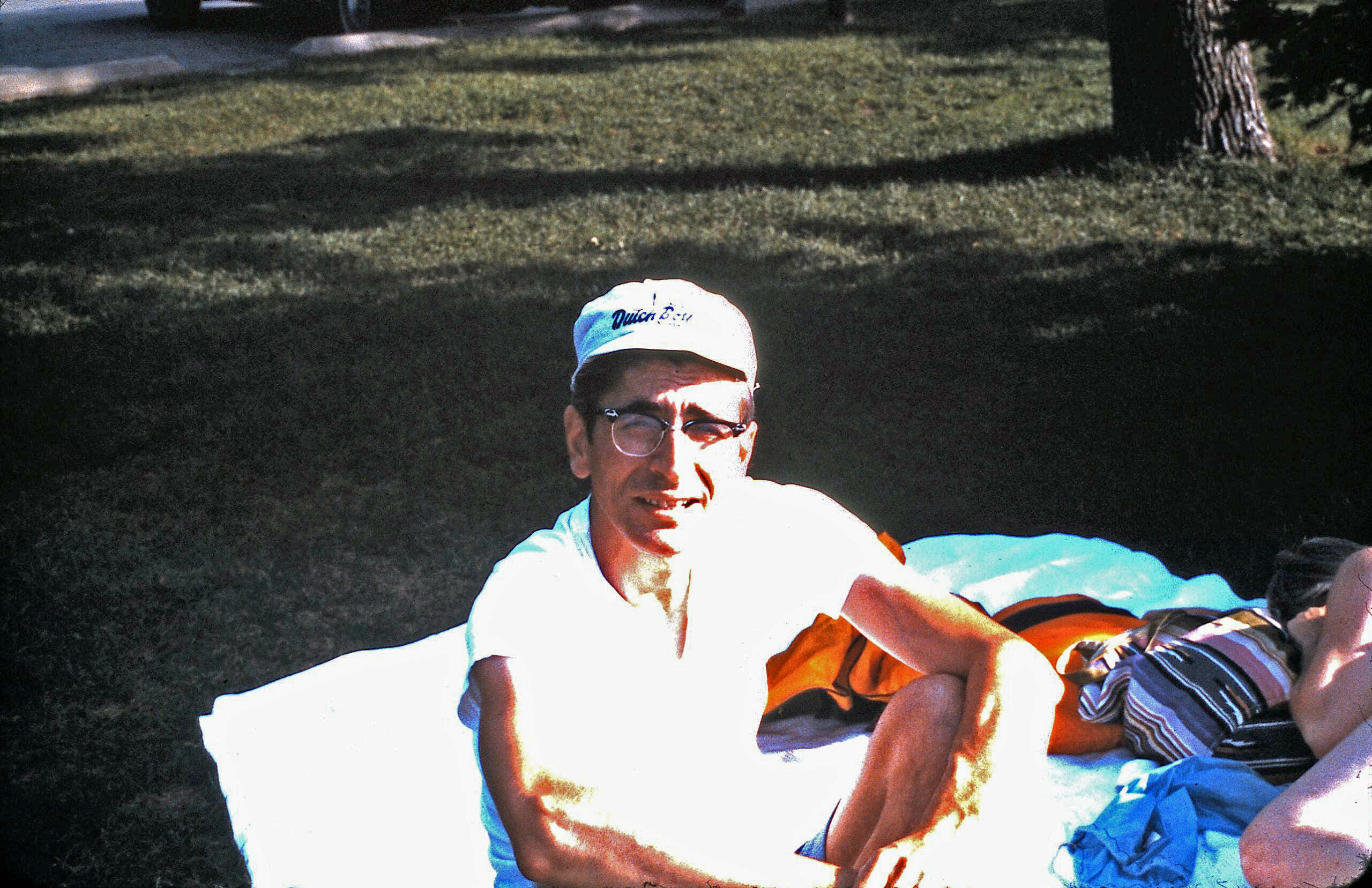 Smiling man wearing a white cap and glasses, outdoors on a sunny day.