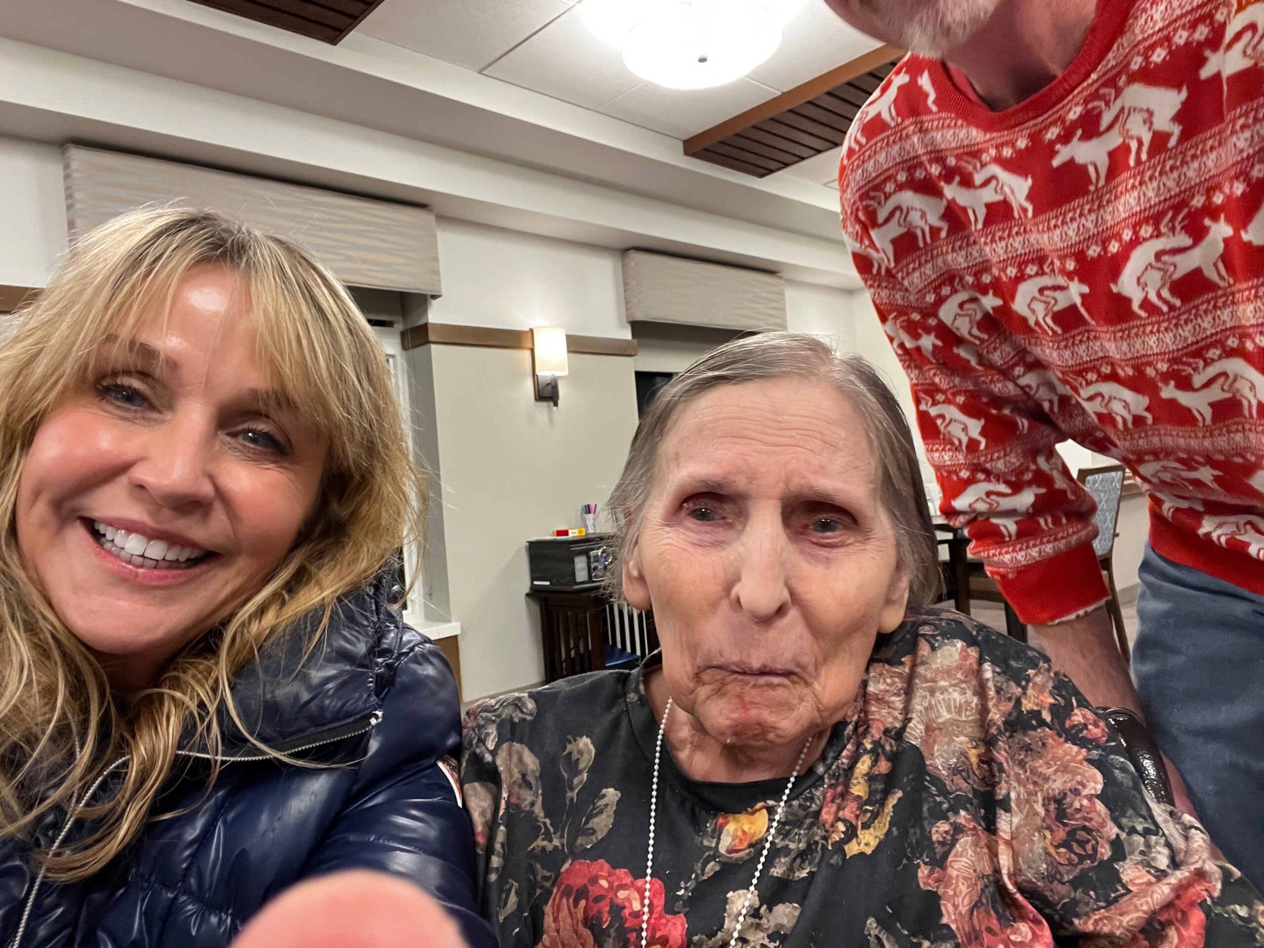 Three people taking a casual indoor selfie: a smiling woman with blonde hair on the left, an older woman in the center, and a person in a bright red patterned sweater on the right.