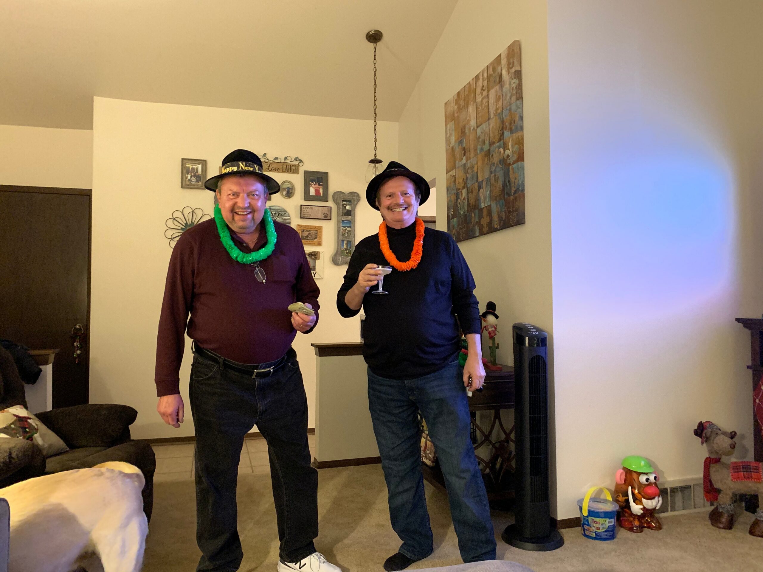 Two men in a home party setting pose for a photo, wearing black hats and bright leis, smiling at the camera.”