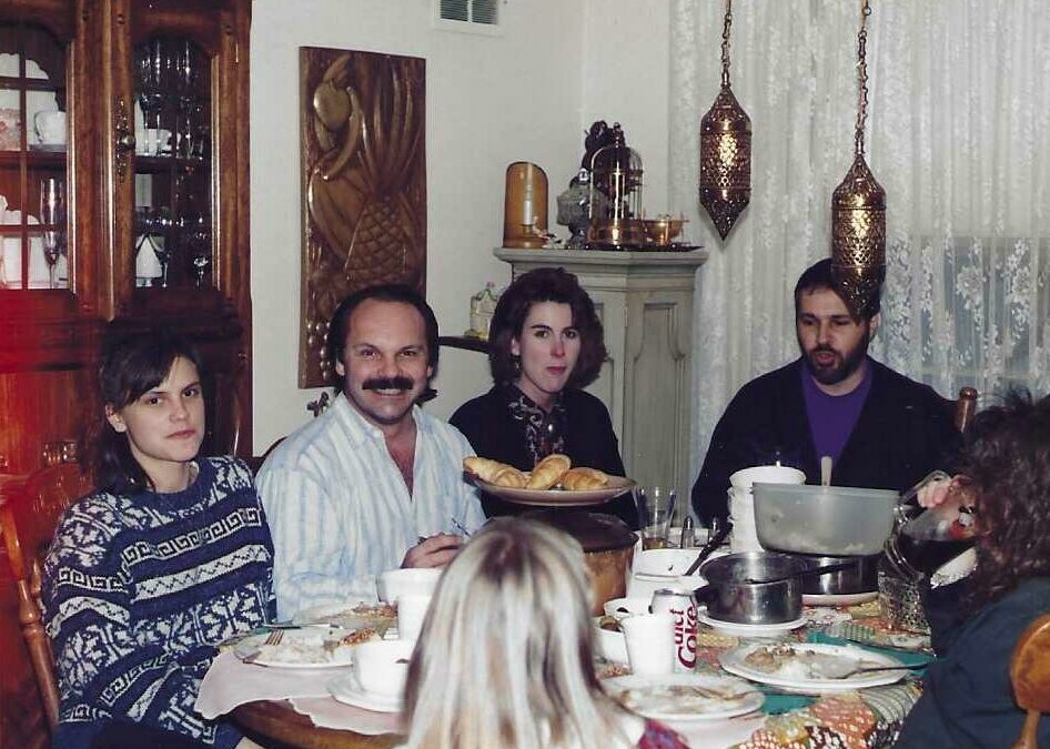 A group of adults sit around a dining table sharing a meal in a cozy, vintage-style room with pendant lamps and a bread basket in the center.
