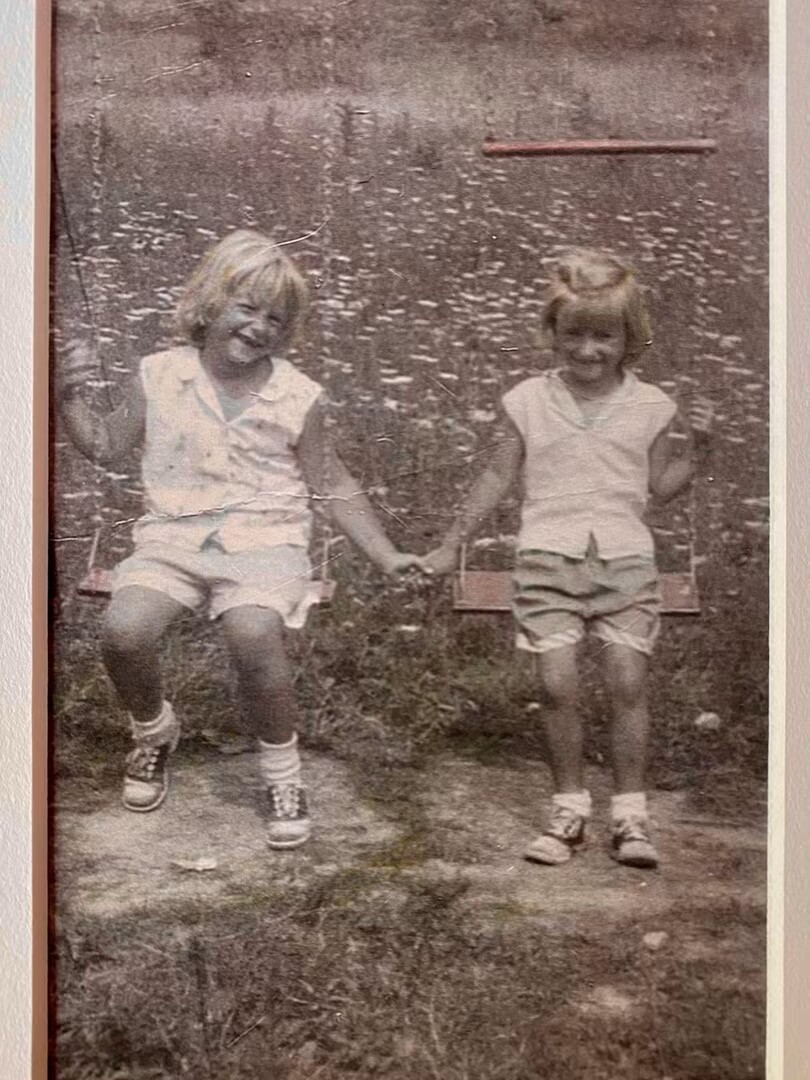 Two young children sit on a swing set in a field, holding hands and smiling at the camera in a vintage sepia-toned photo.