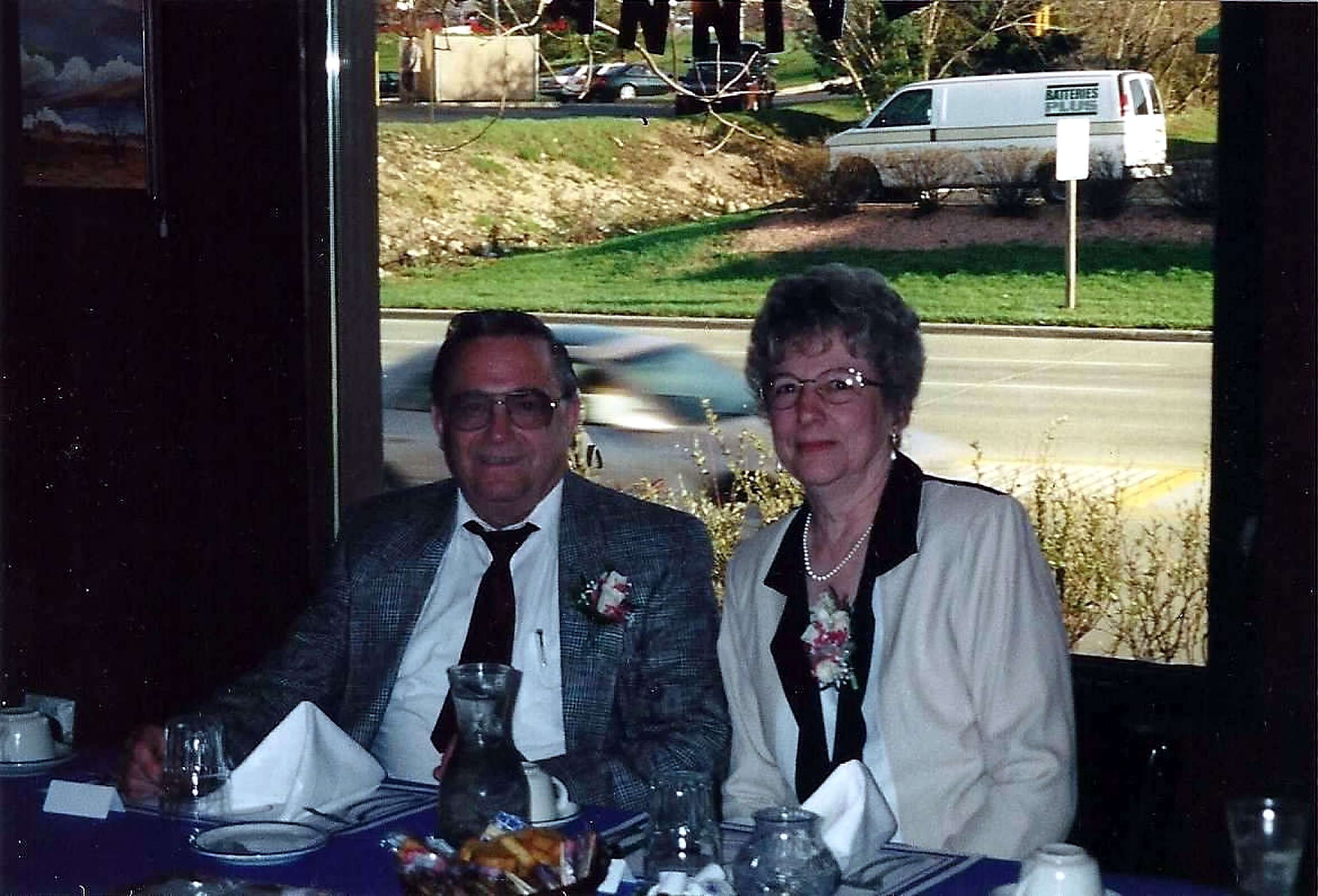 A smiling couple in formal attire sit at a table with drinks and flowers during a celebration, indoors by a window.