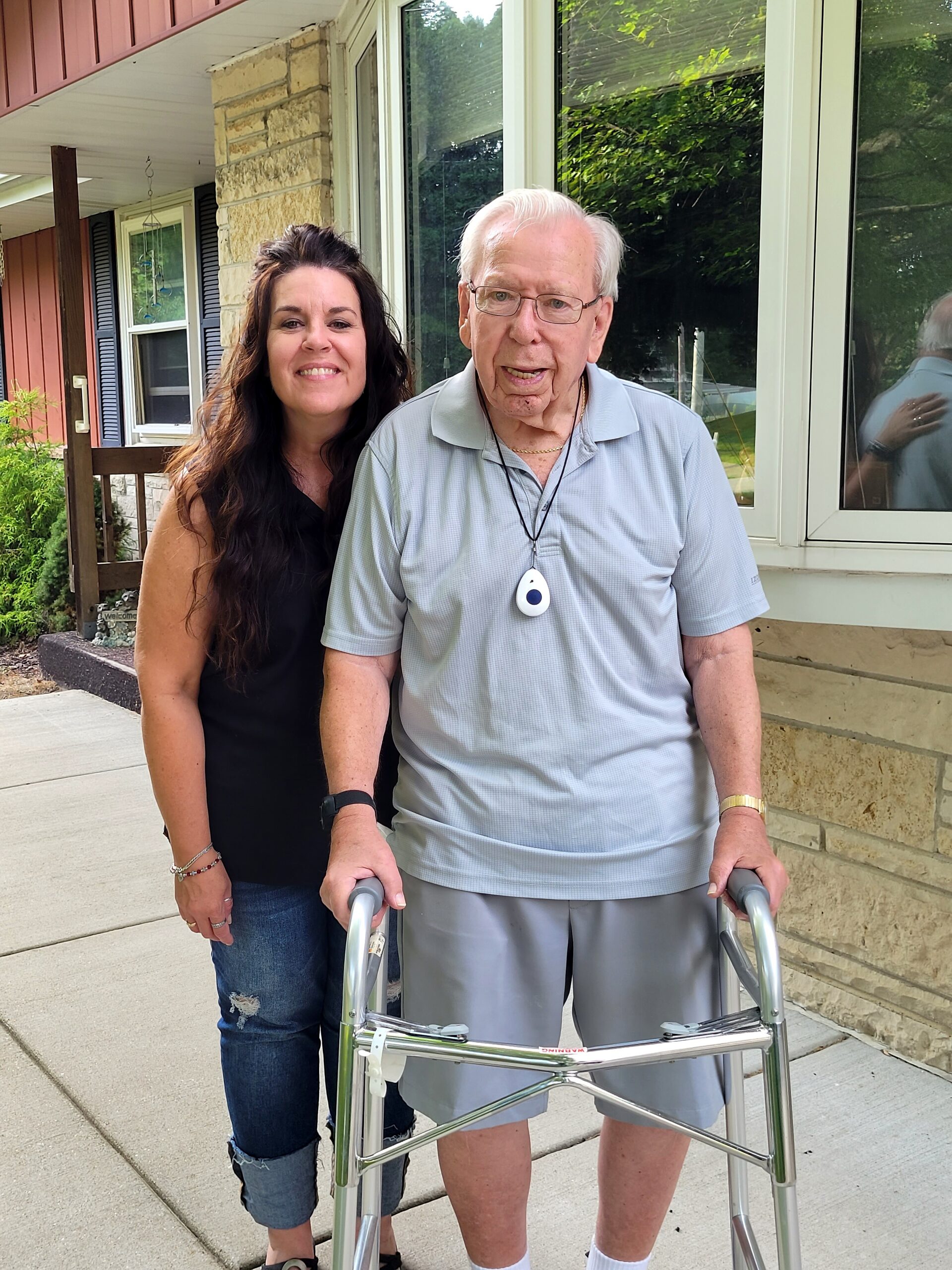 Woman with long dark hair standing beside an elderly man using a walker on a porch outside a house.