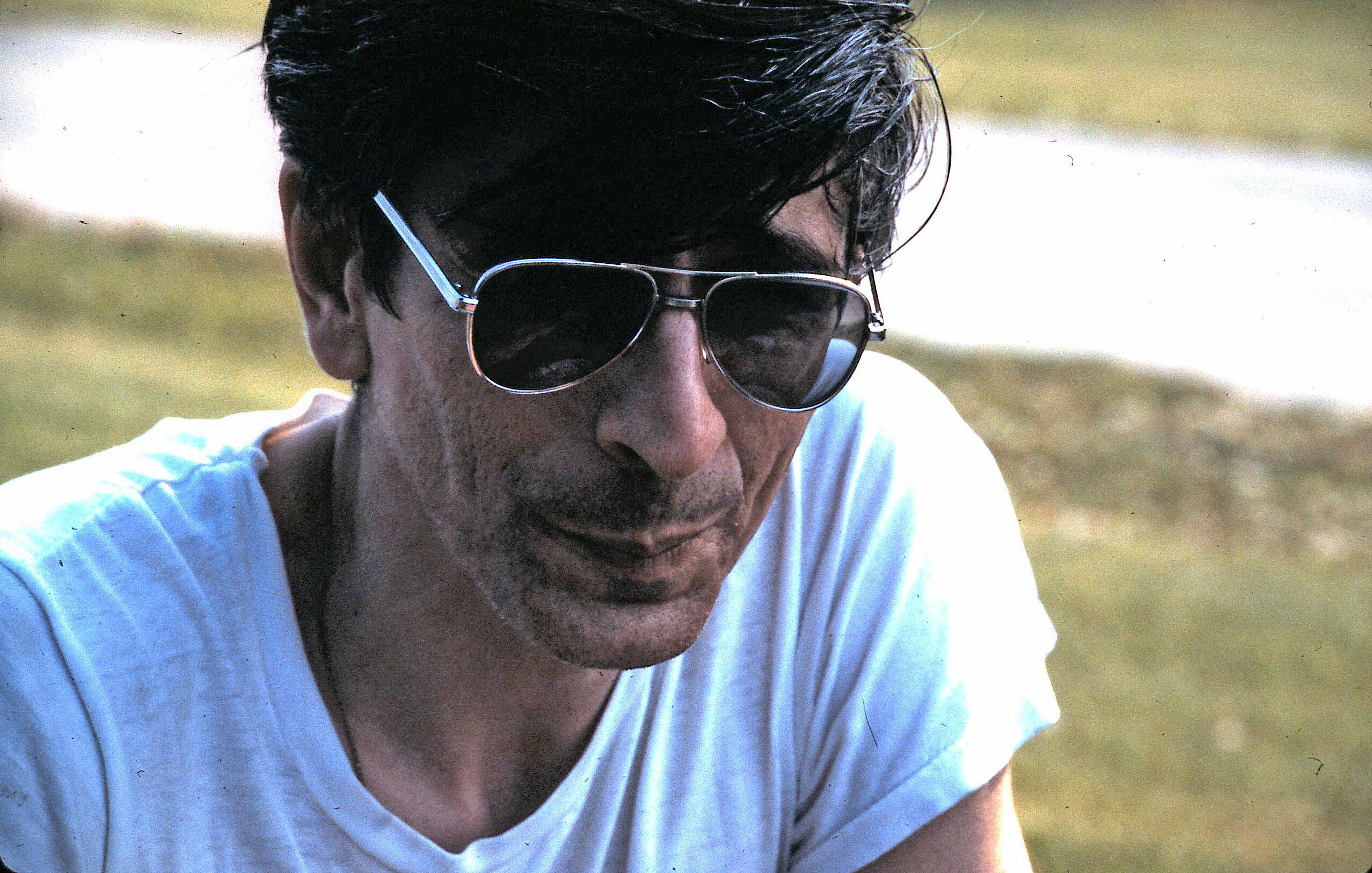 Close-up of a man wearing aviator sunglasses and a white T-shirt, looking downward in a sunny outdoor setting.