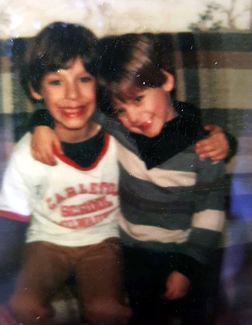 Two young boys sit close together with arms around each other, smiling at the camera.
