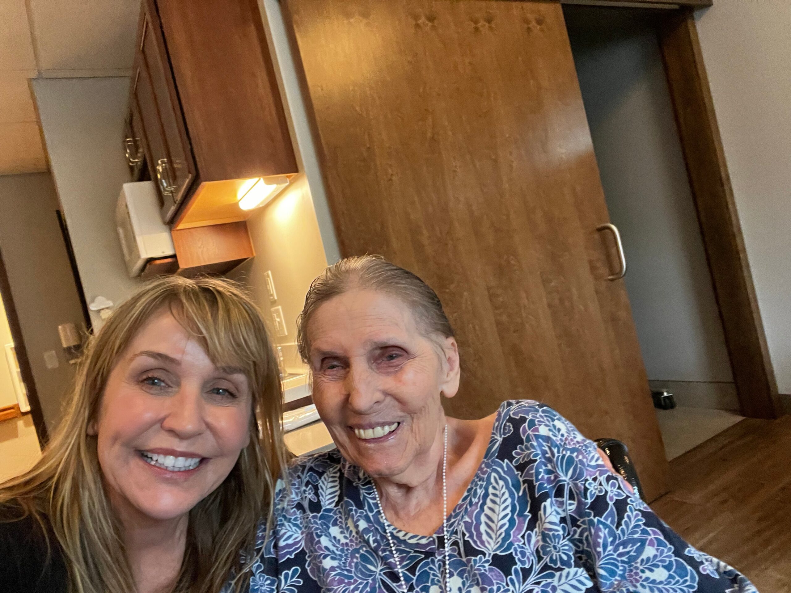 Two women posing for a selfie indoors, both smiling at the camera in a wood-toned room with cabinets behind them.