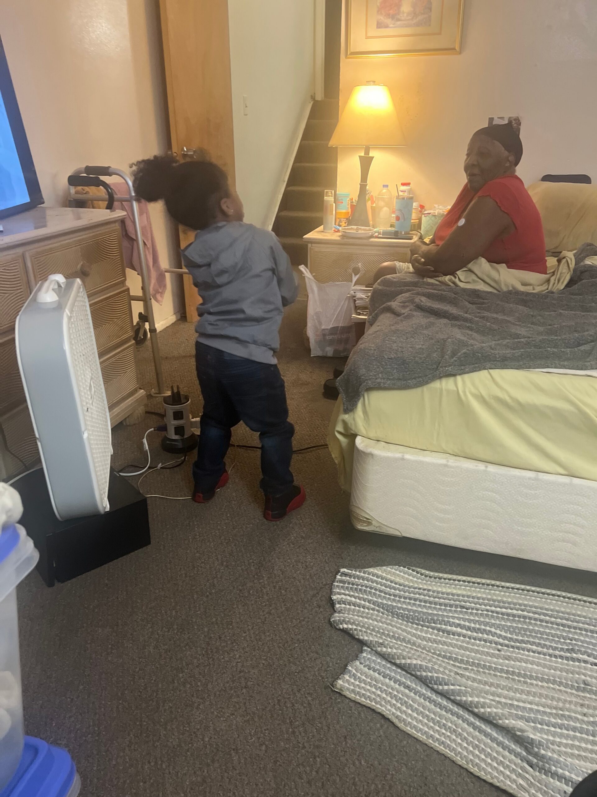 Young child facing a seated elderly person in bed, surrounded by clutter and a lamp in a cozy bedroom.