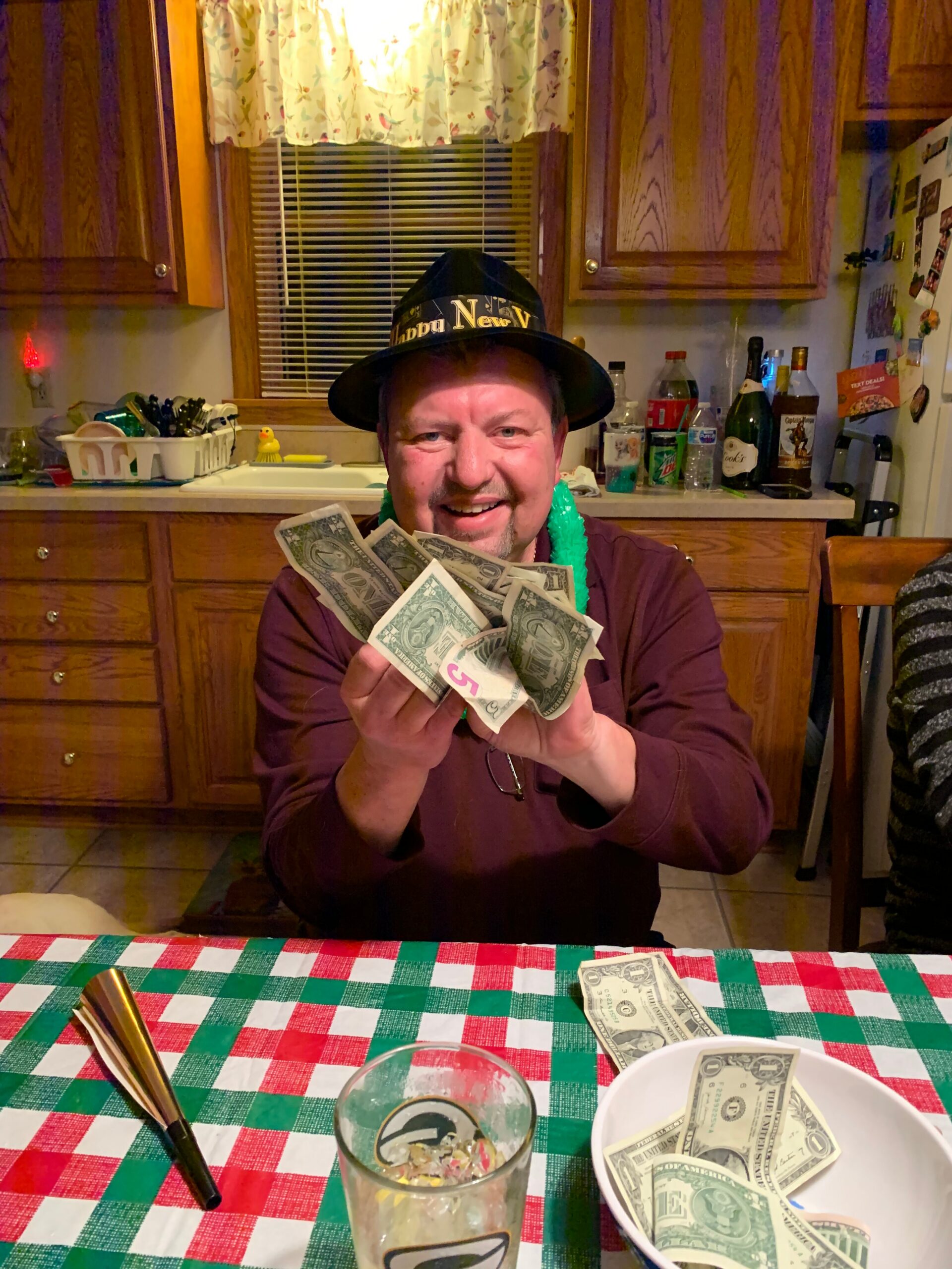 Smiling man wearing a festive hat that says 'Happy New Year' holds fan of dollar bills at a kitchen table with a red-and-green checkered tablecloth in the foreground.