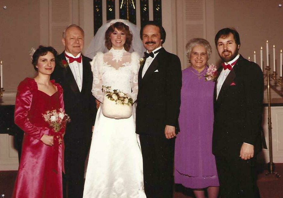 Wedding party: bride in white gown with bouquet flanked by family members in formal attire inside a church setting.