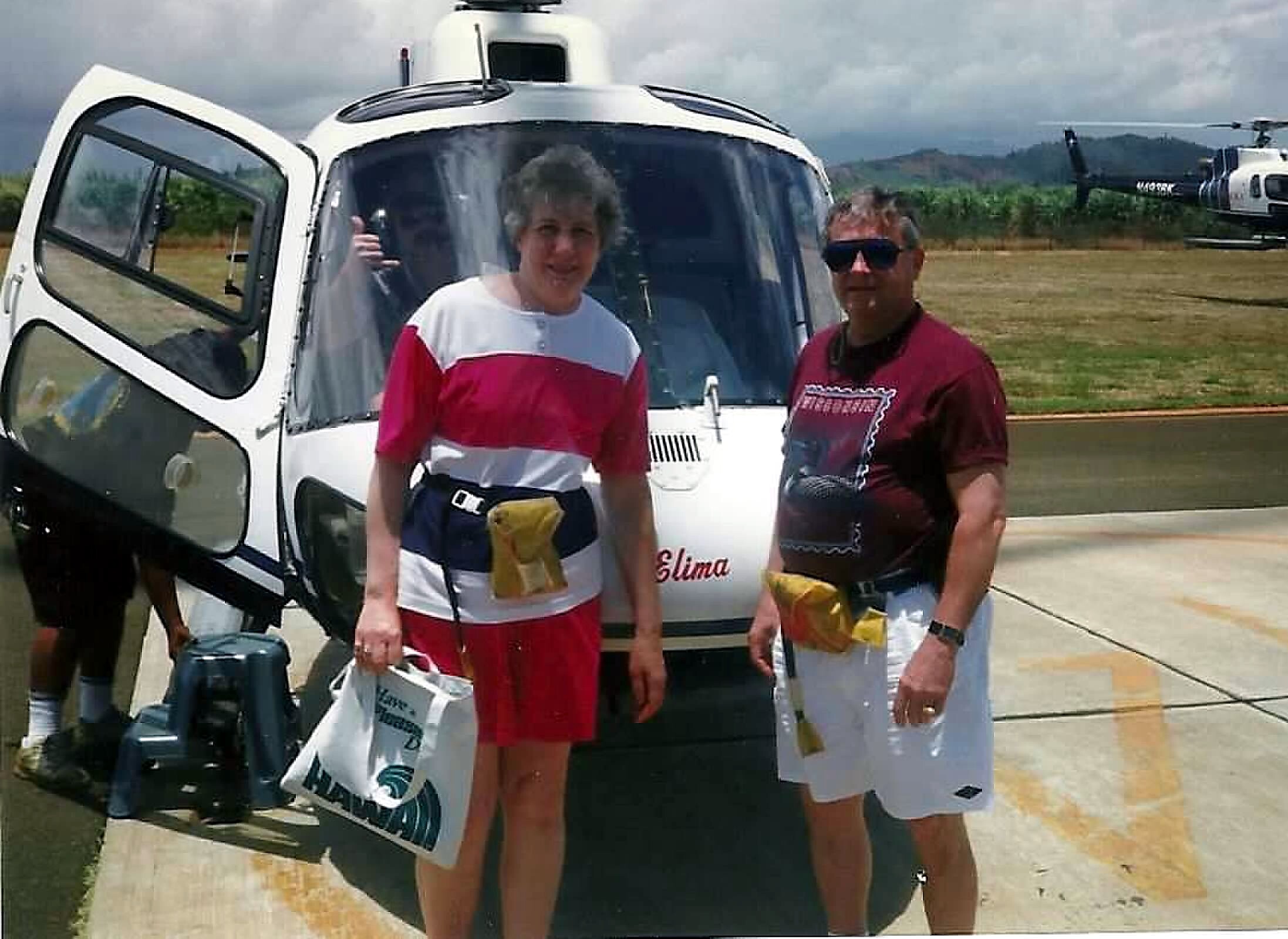 Two adults posing beside a white helicopter on a paved airfield, holding bags.