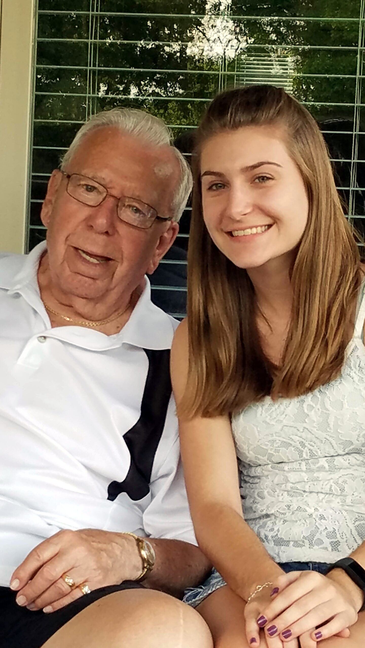 Grandfather and granddaughter smiling while sitting together on a porch with blinds in the background.