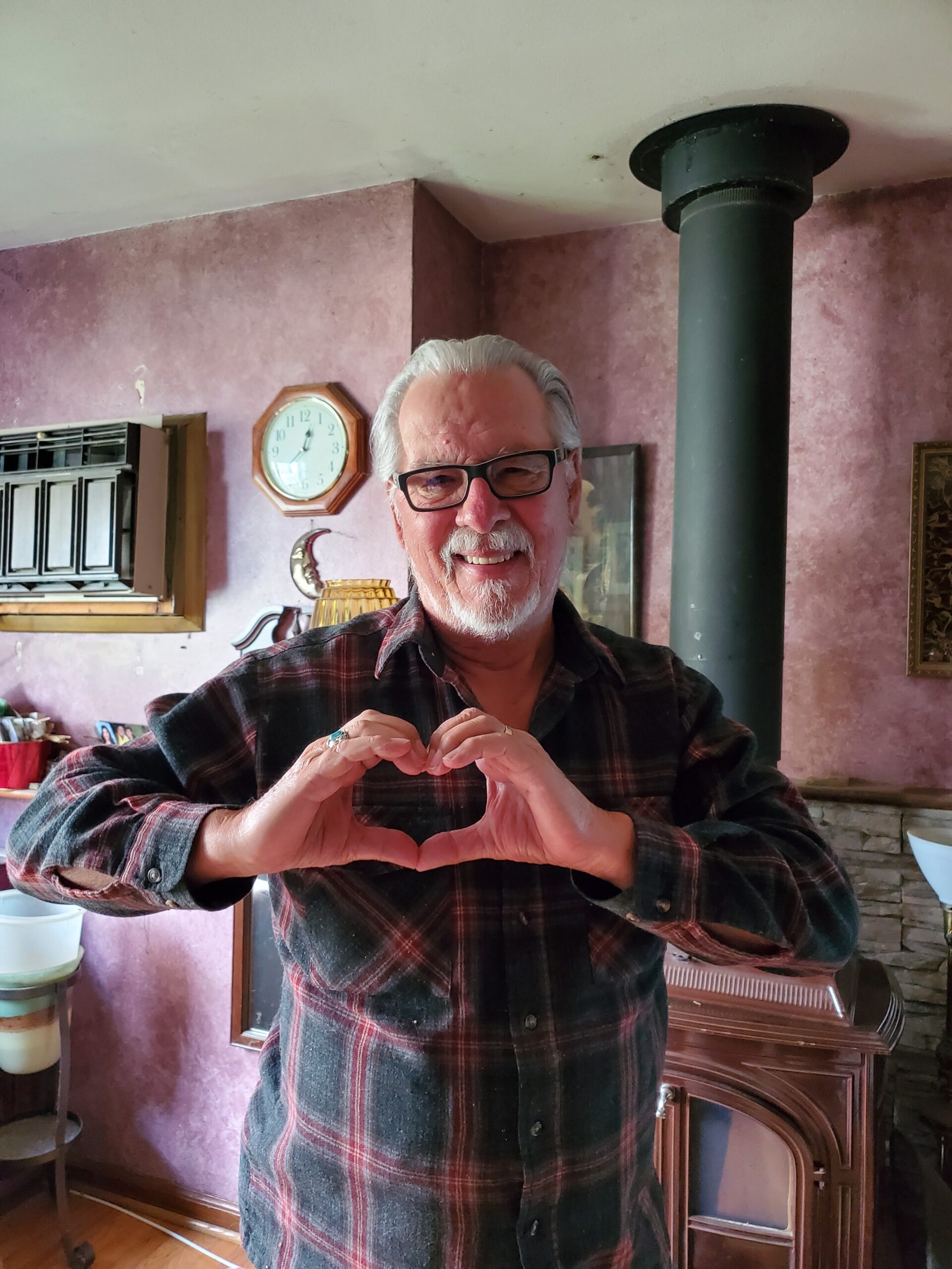 Smiling older man with glasses making a heart shape with his hands in a cozy, pink-walled room.